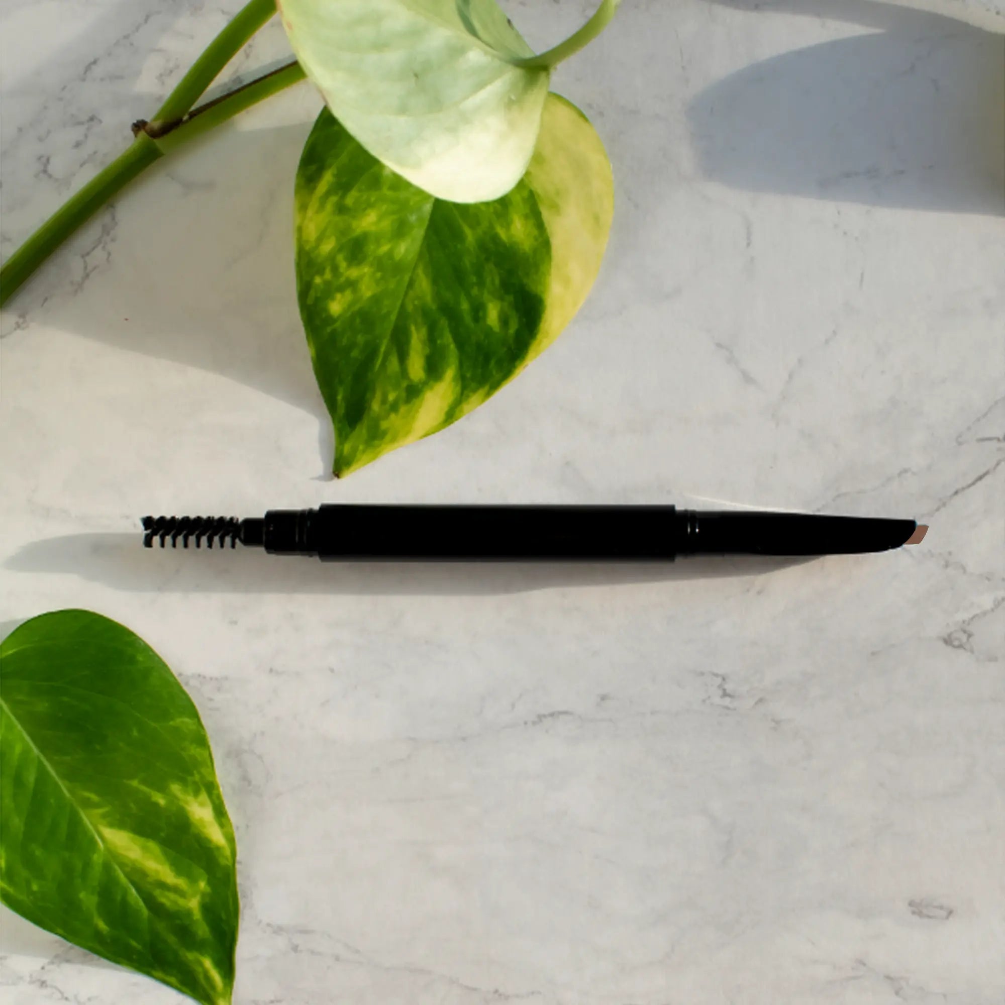 Automatic eyebrow pencil in taupe with a spoolie brush on a marble surface with leaves.