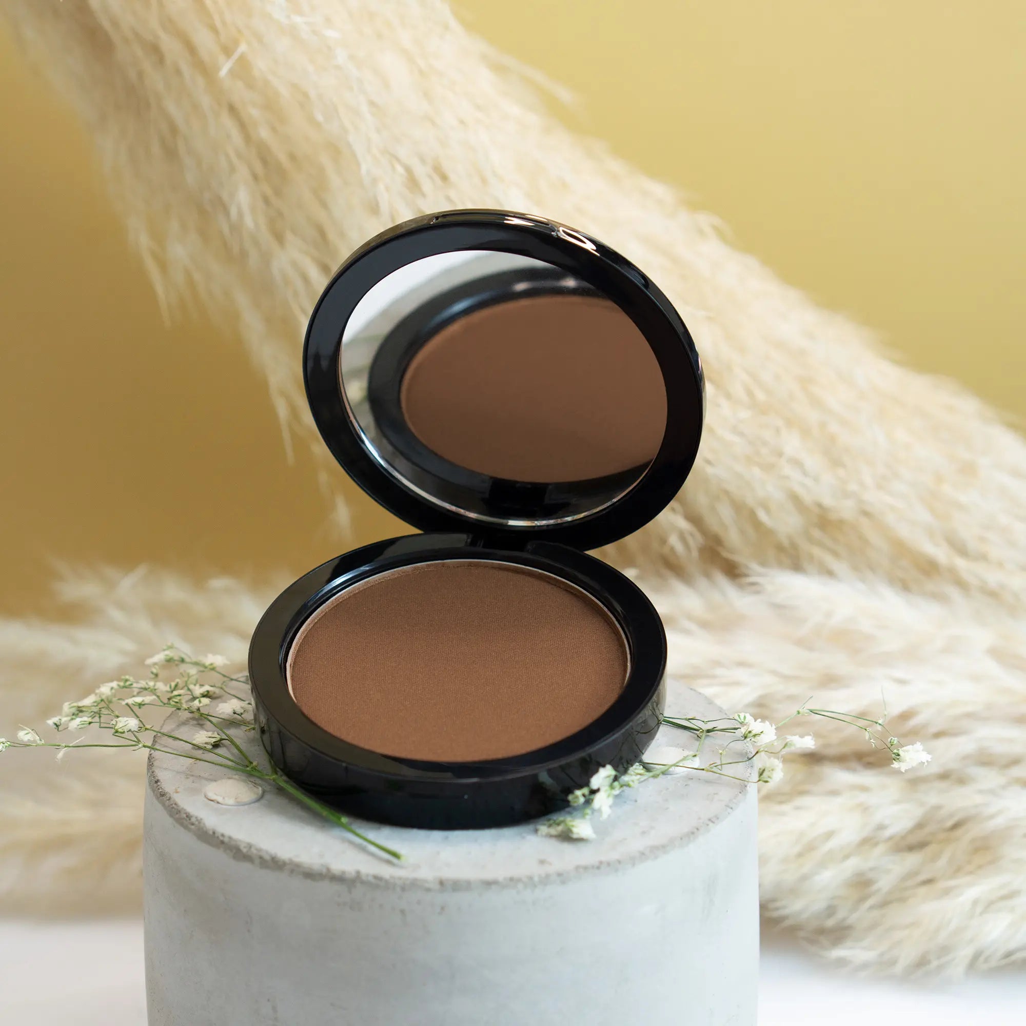 Open compact bronzer in Mocha with a mirror, on a concrete stand with white flowers.