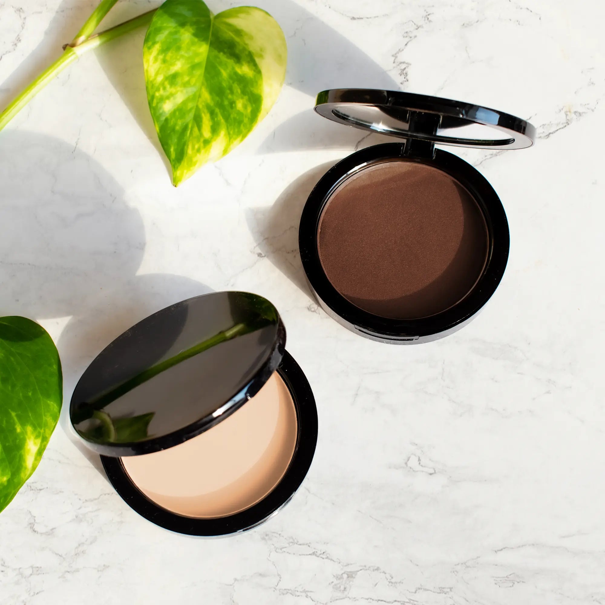 Two open compacts of powder foundation, one light and one dark, on a marble surface.