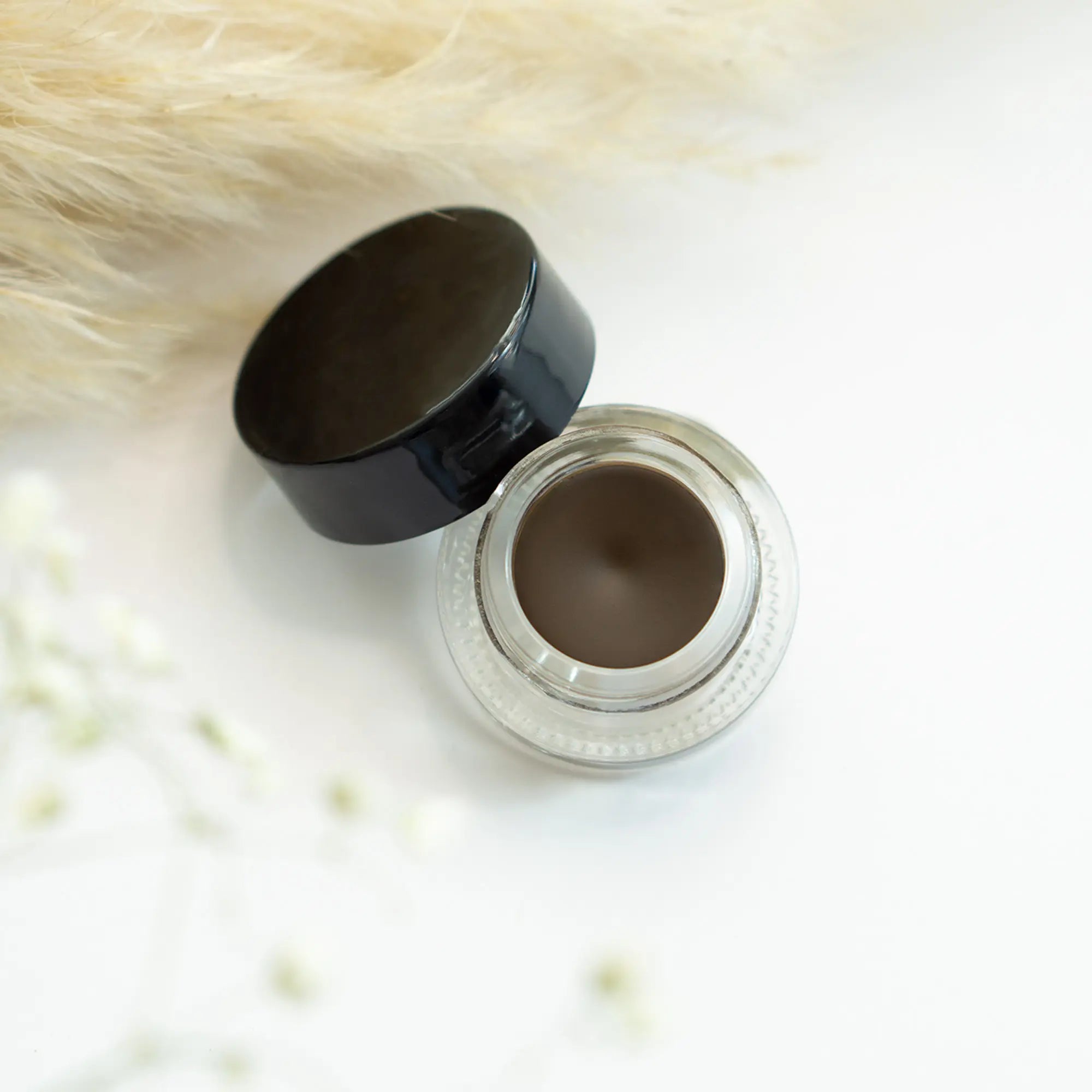 An open jar of brown gel eyeliner with a black cap, on a white surface with feathers.