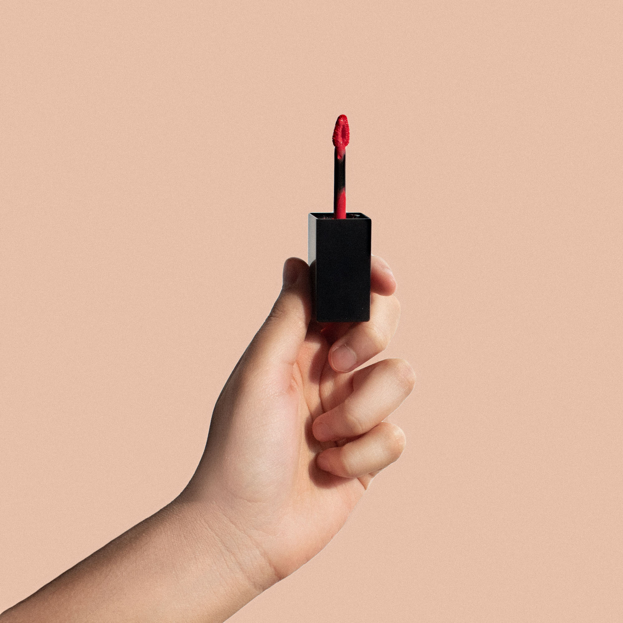 A hand holds a matte lip stain with a dark cherry red applicator against a peach background.