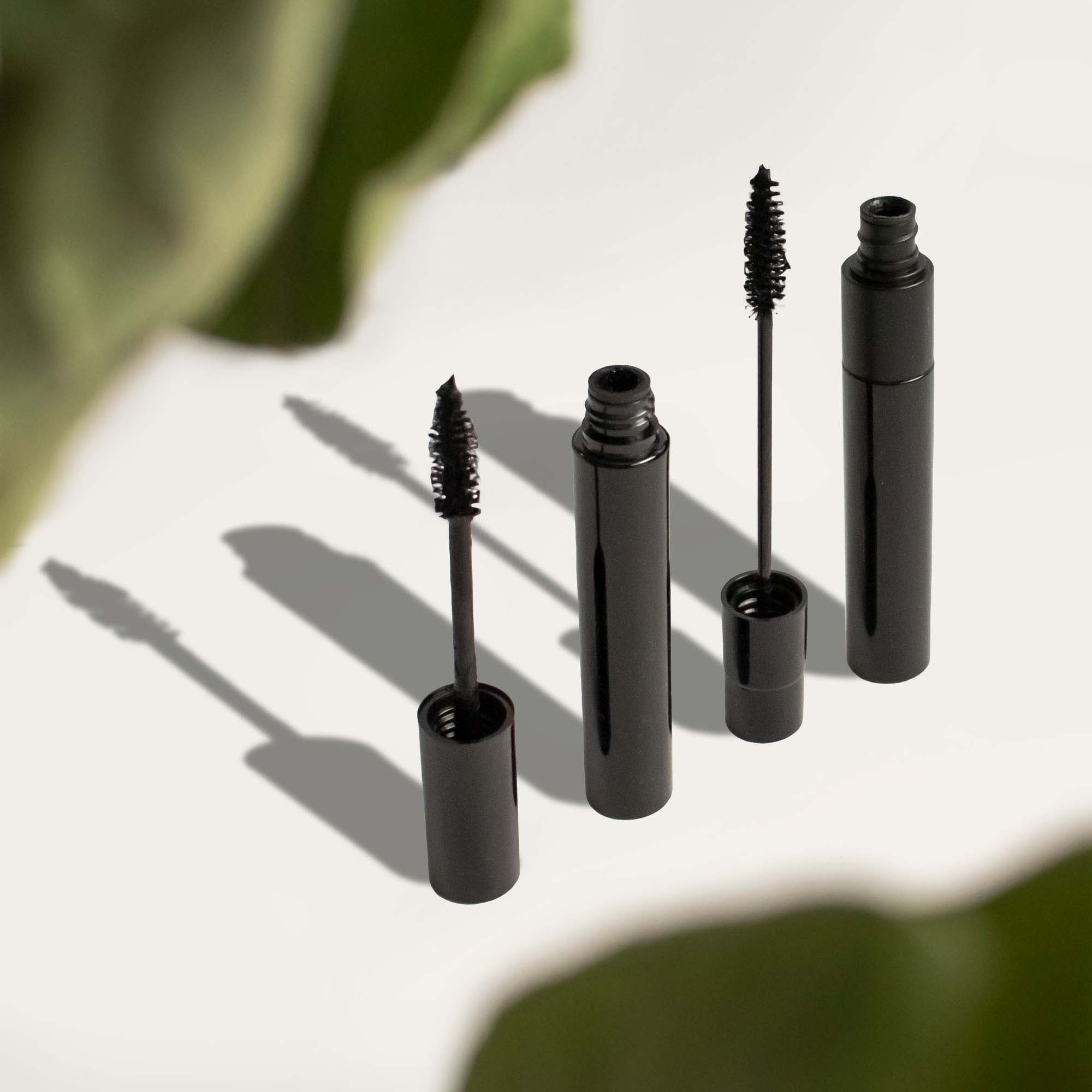 Two black mascara tubes with wands are displayed on a white surface with green leaves.