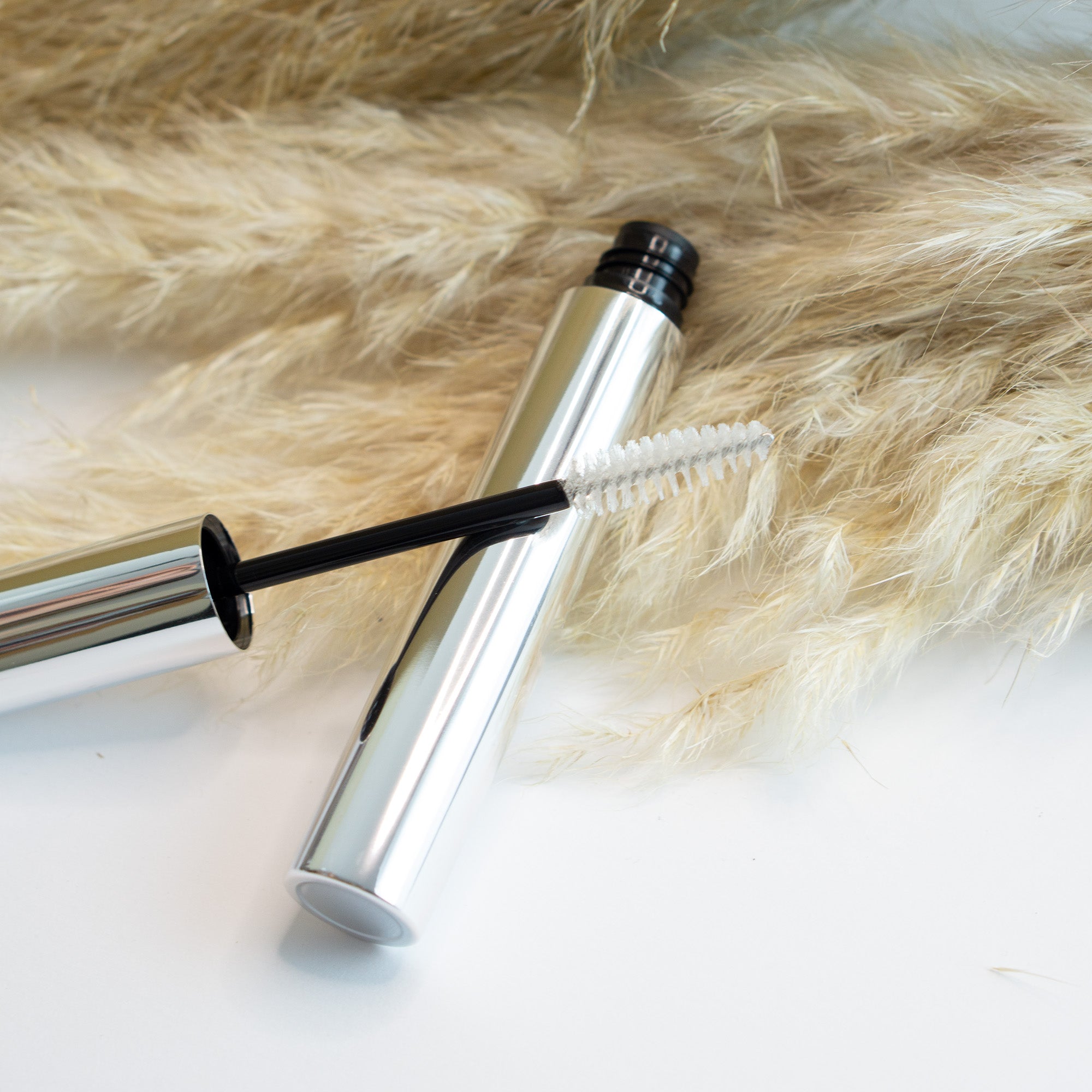 Clear eyebrow gel with an applicator brush rests on a white surface with dried pampas grass.