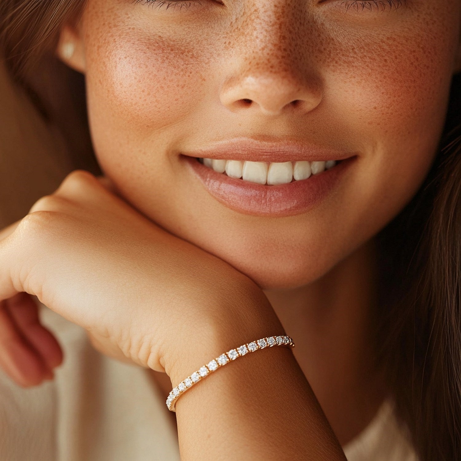 A woman with freckles smiles, resting her chin on her hand, wearing a yellow gold diamond tennis bracelet.
