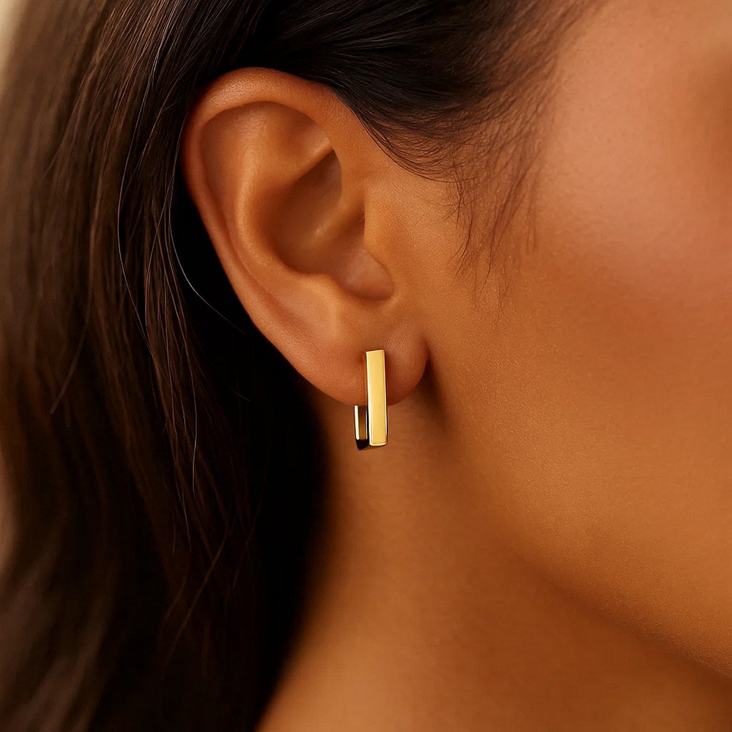 A woman wears a 14k yellow gold rectangle huggie hoop earring.