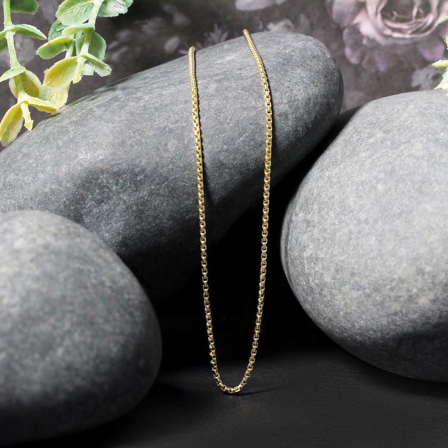 A 14k yellow gold round box chain is draped over smooth gray stones.