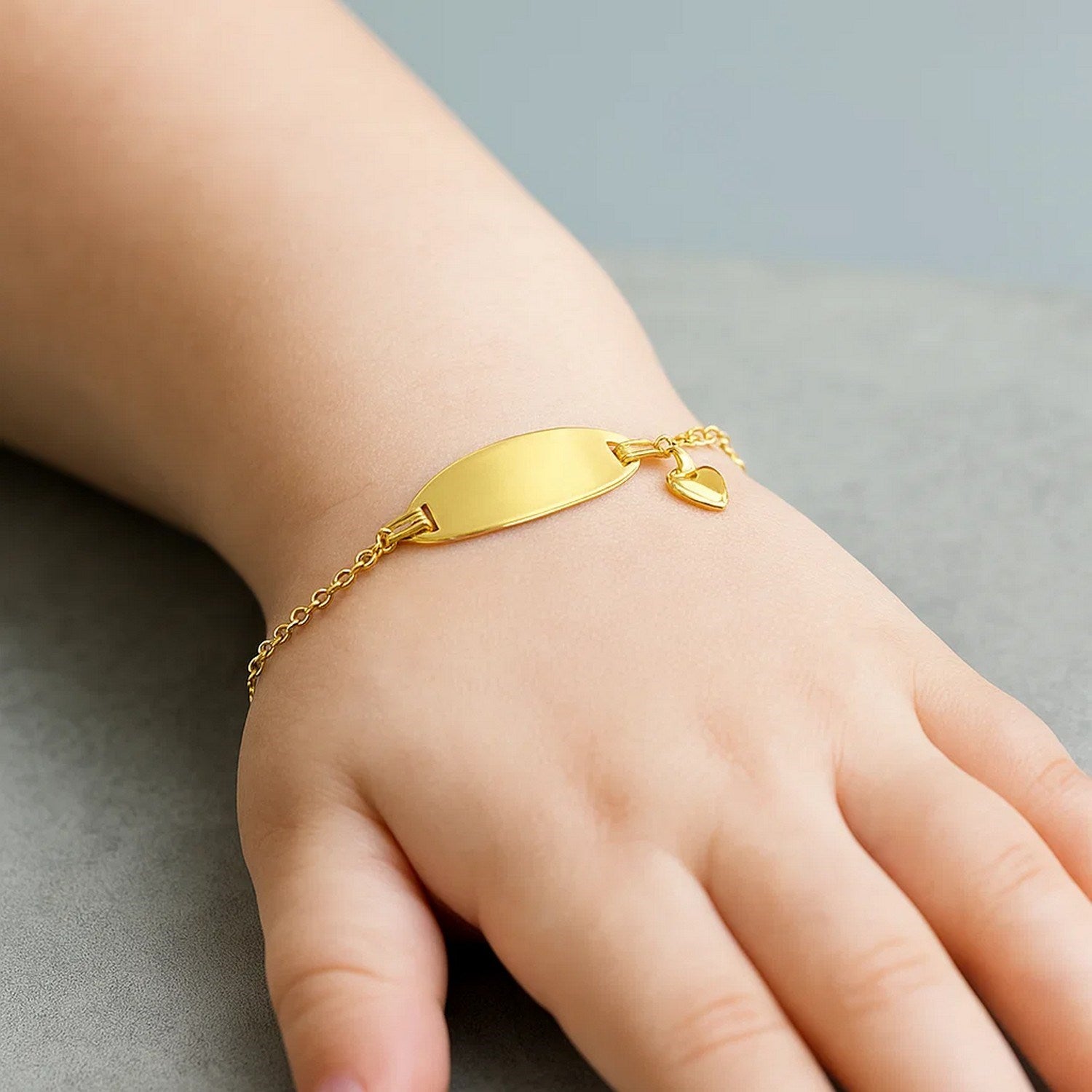 A child's hand wears a 14k yellow gold ID bracelet with a heart charm.