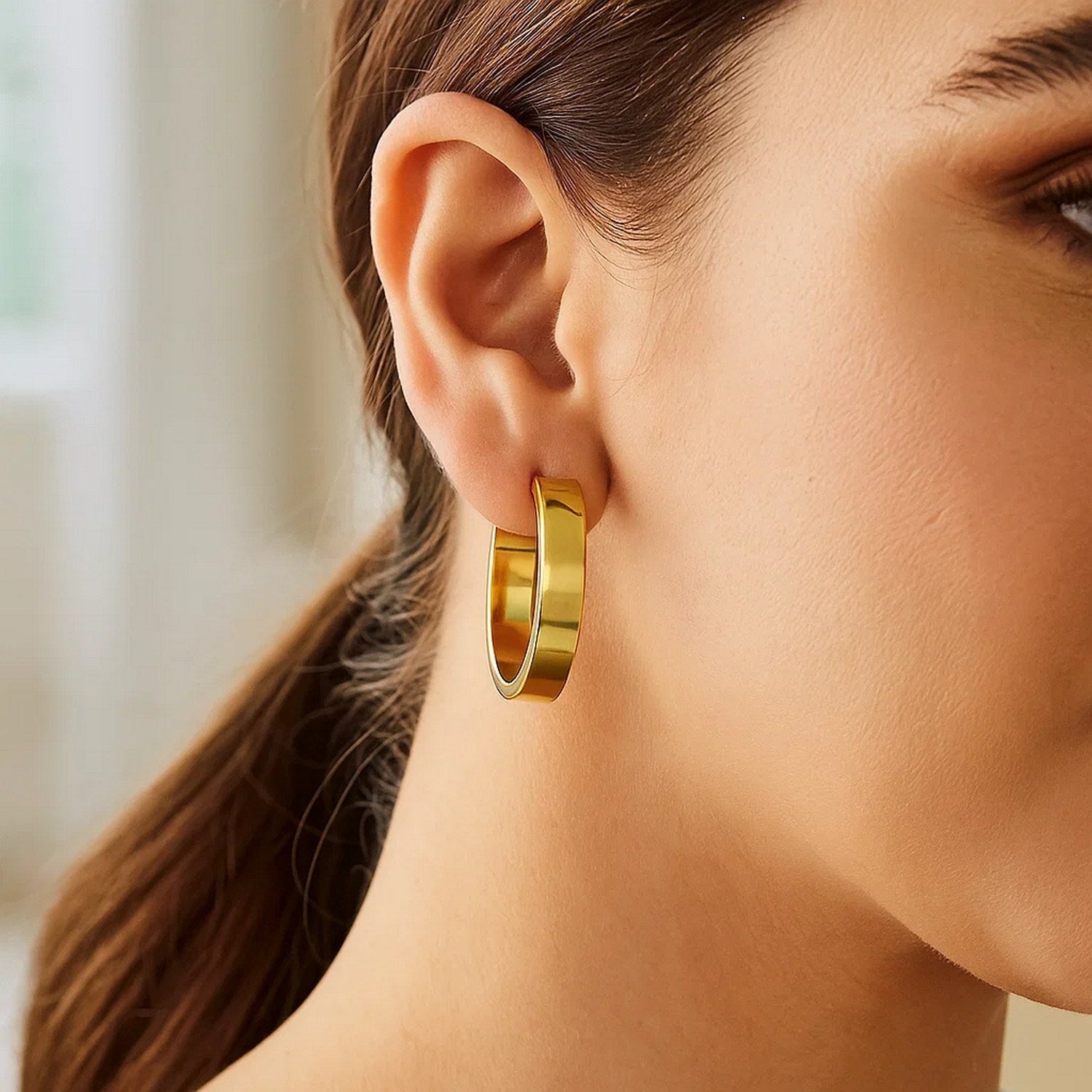 A woman wears a single medium tube-shaped hoop earring in 14K yellow gold.