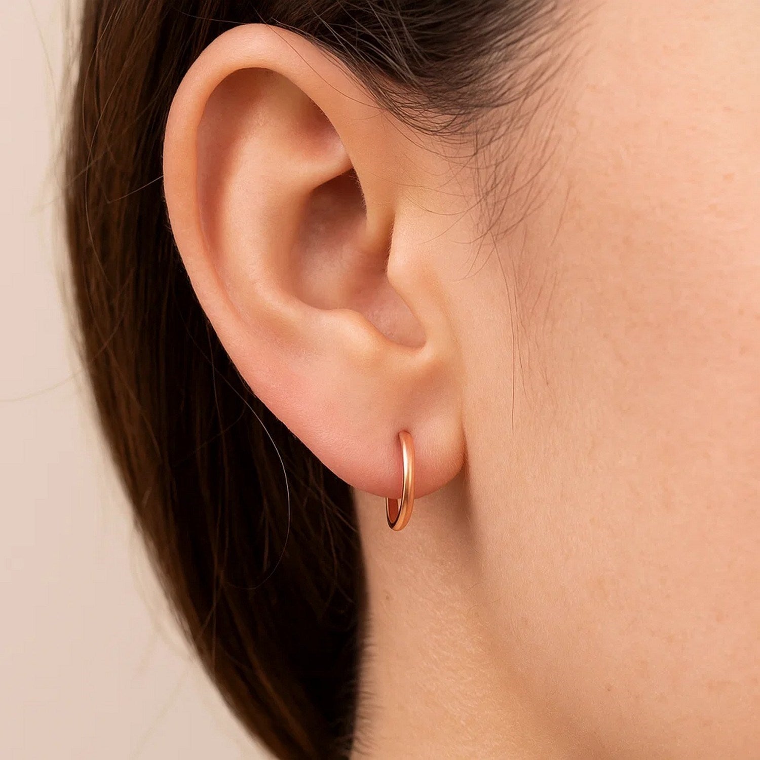 A single ear wears a petite, polished 14k rose gold hoop earring.