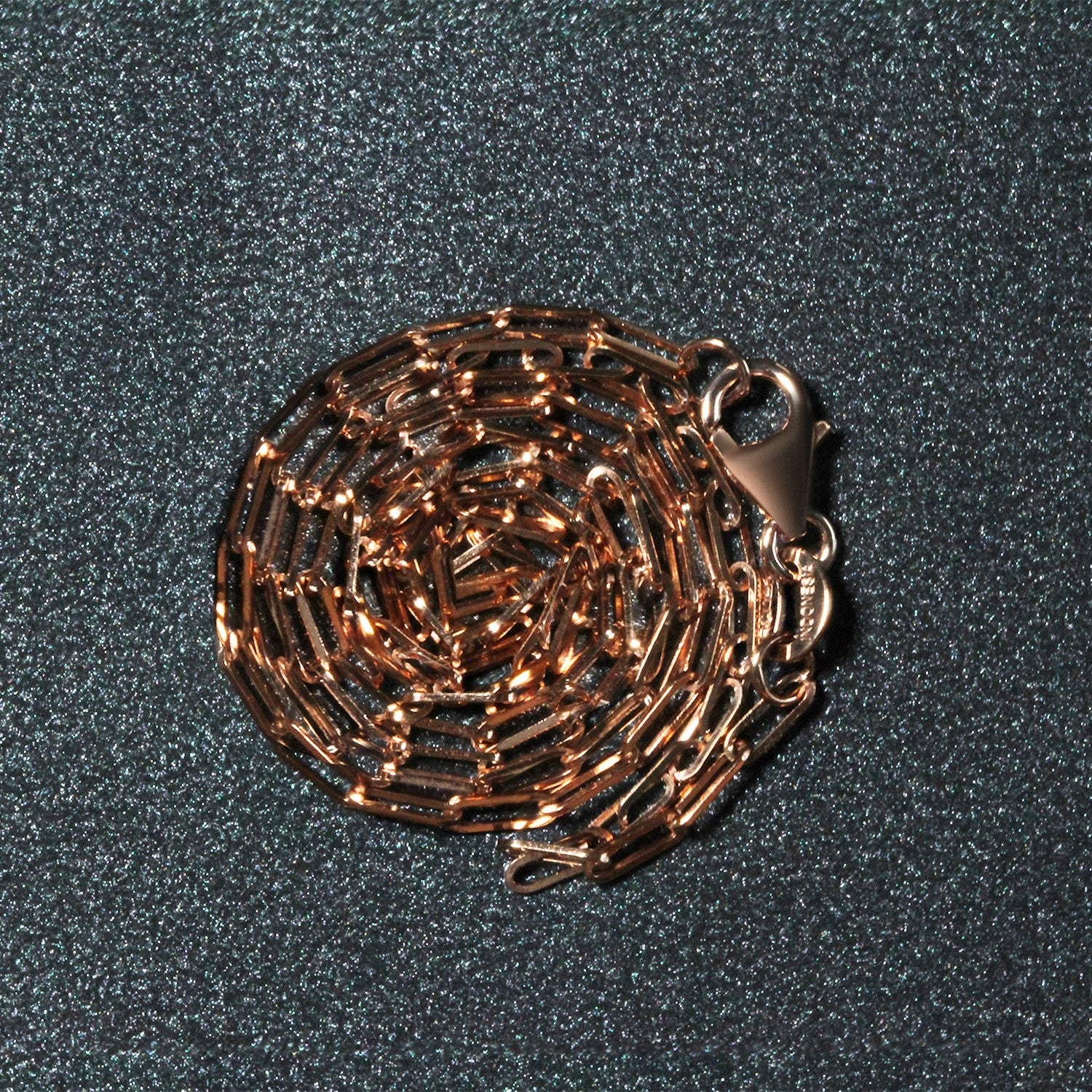 14K rose gold paperclip chain with a lobster clasp, coiled on a dark, glittery surface.