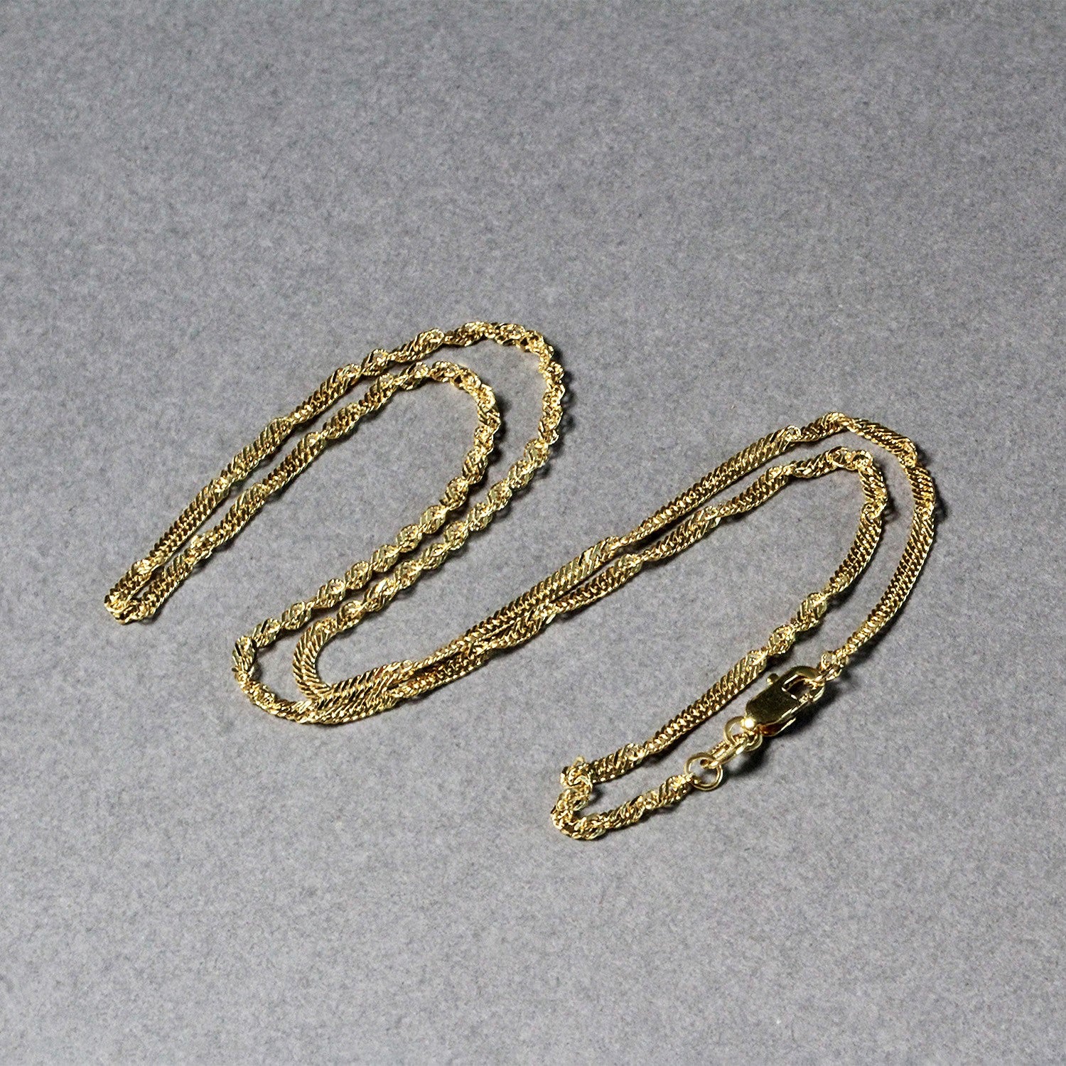 A 14k yellow gold Singapore chain with a lobster clasp lies on a gray surface.