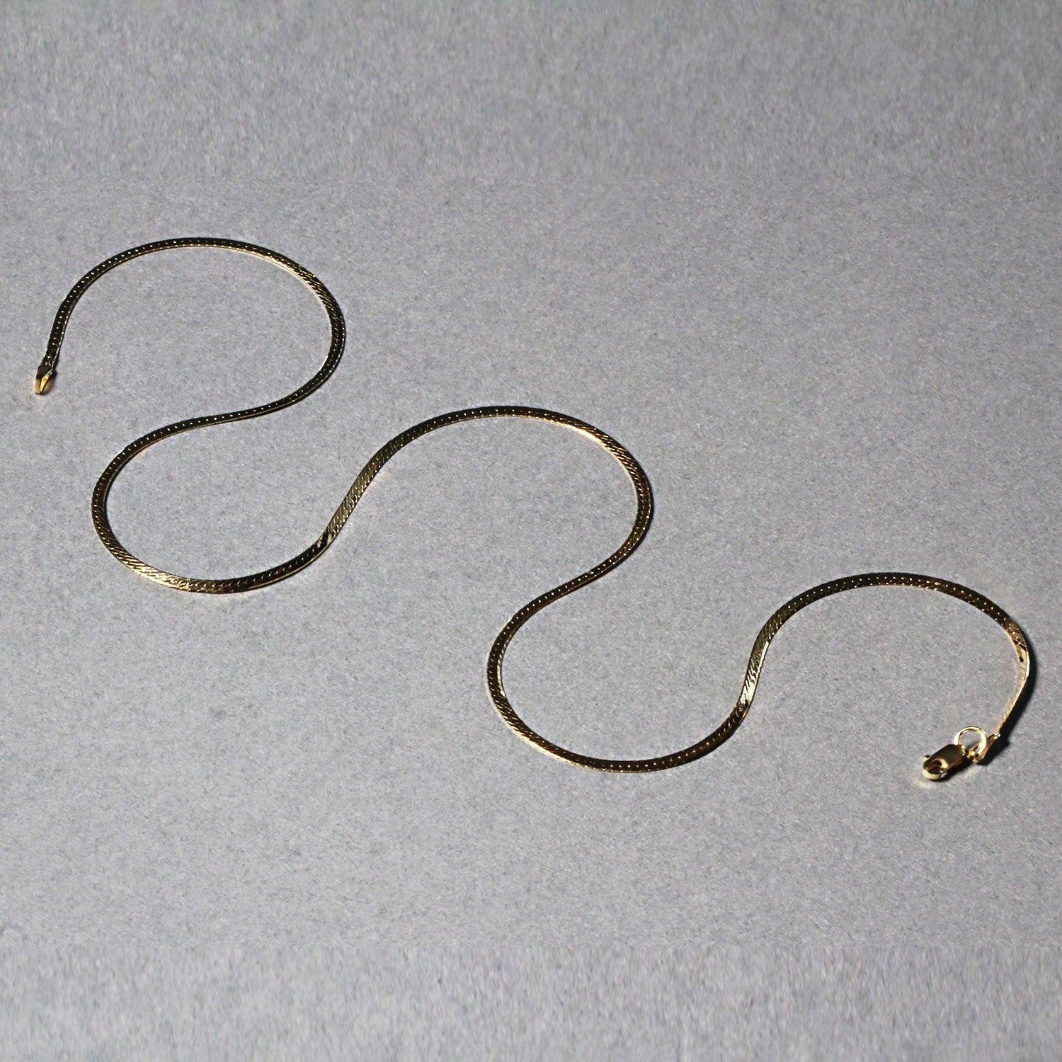 A 14k yellow gold super flex herringbone chain is laid out on a gray surface.
