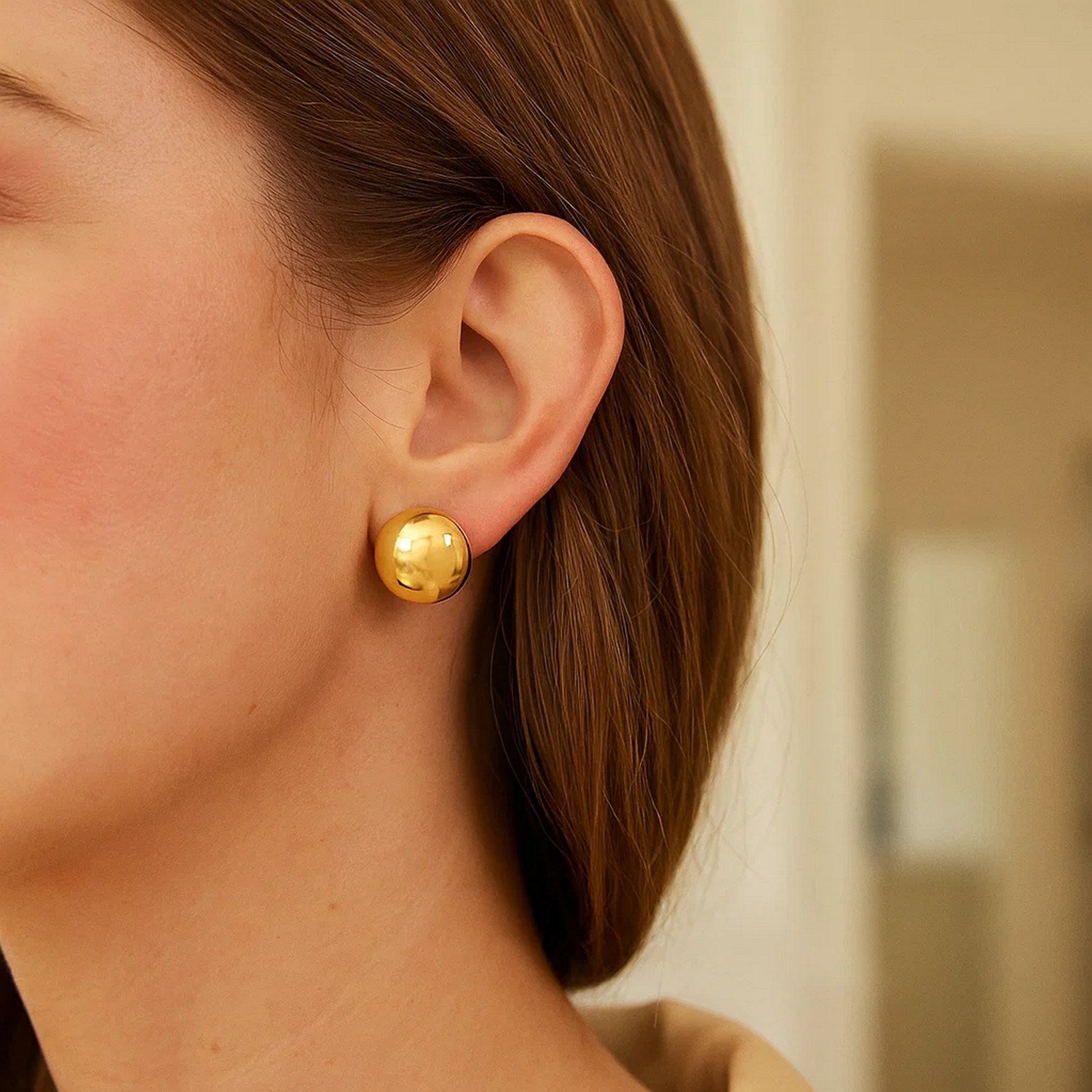 A woman wears a single large, round, polished gold stud earring on her right ear.