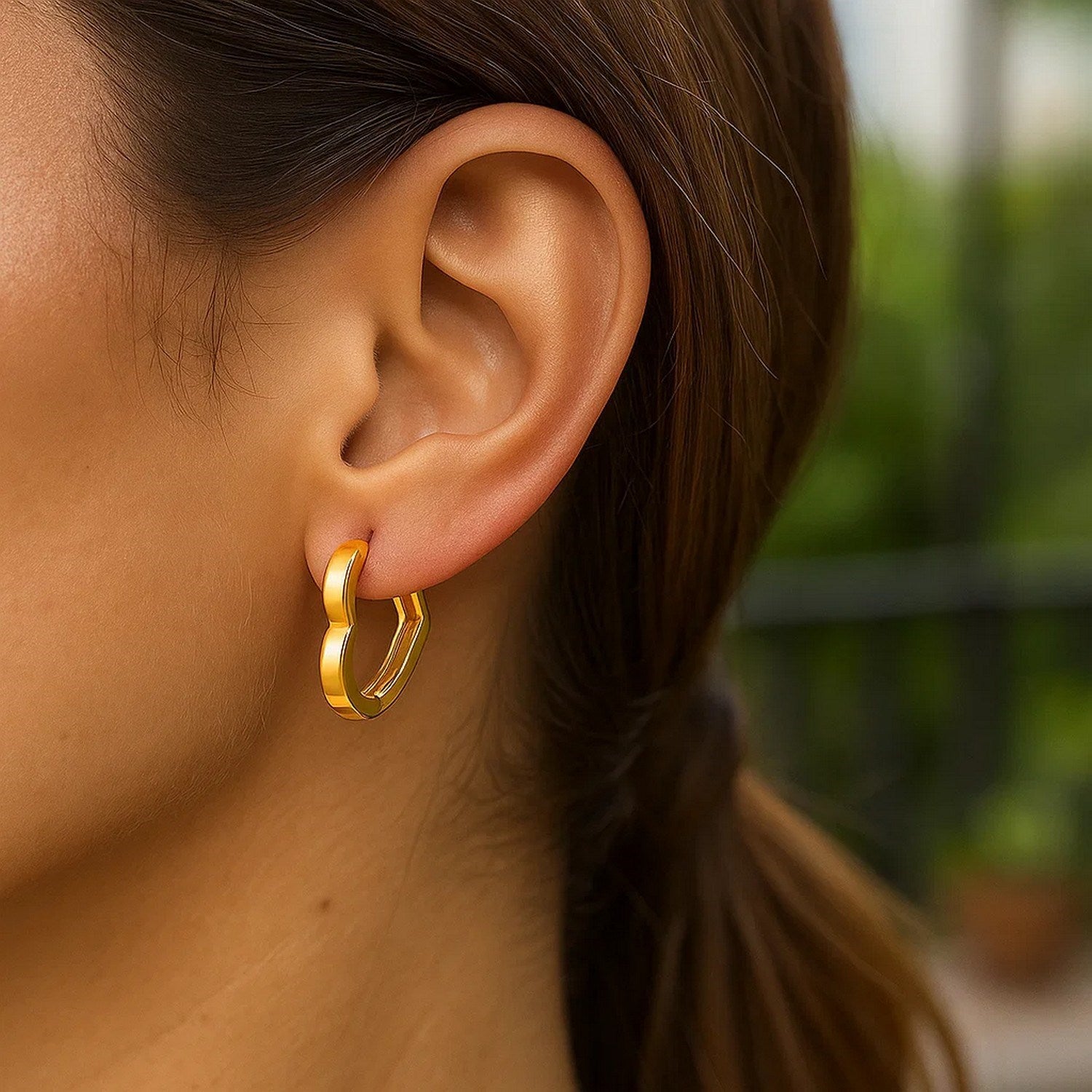 A woman wears a 14k yellow gold heart huggie hoop earring.