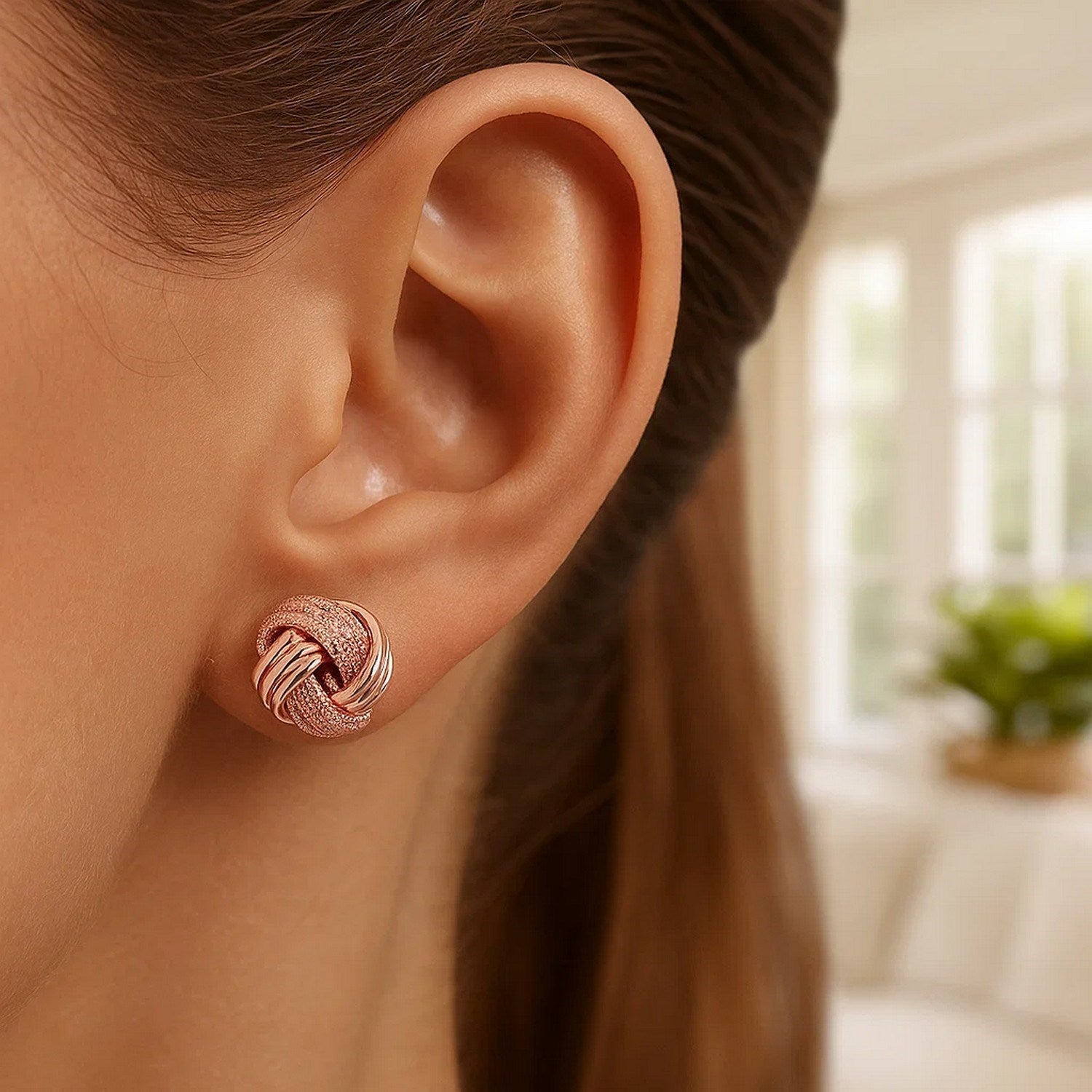 A rose gold love knot earring with ridge texture is worn on an ear.