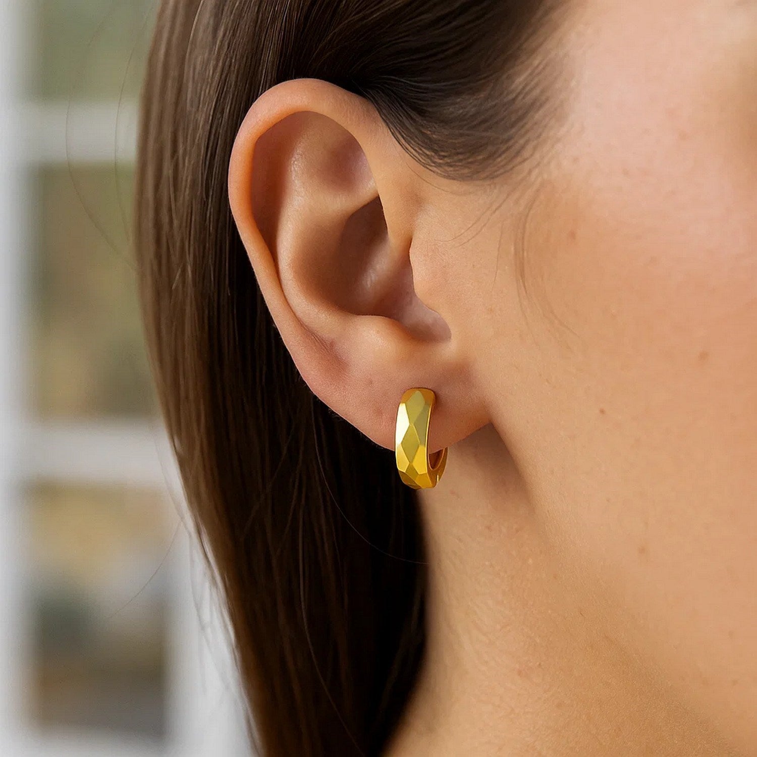 A woman wears a single 14K yellow gold diamond motif faceted huggie earring.