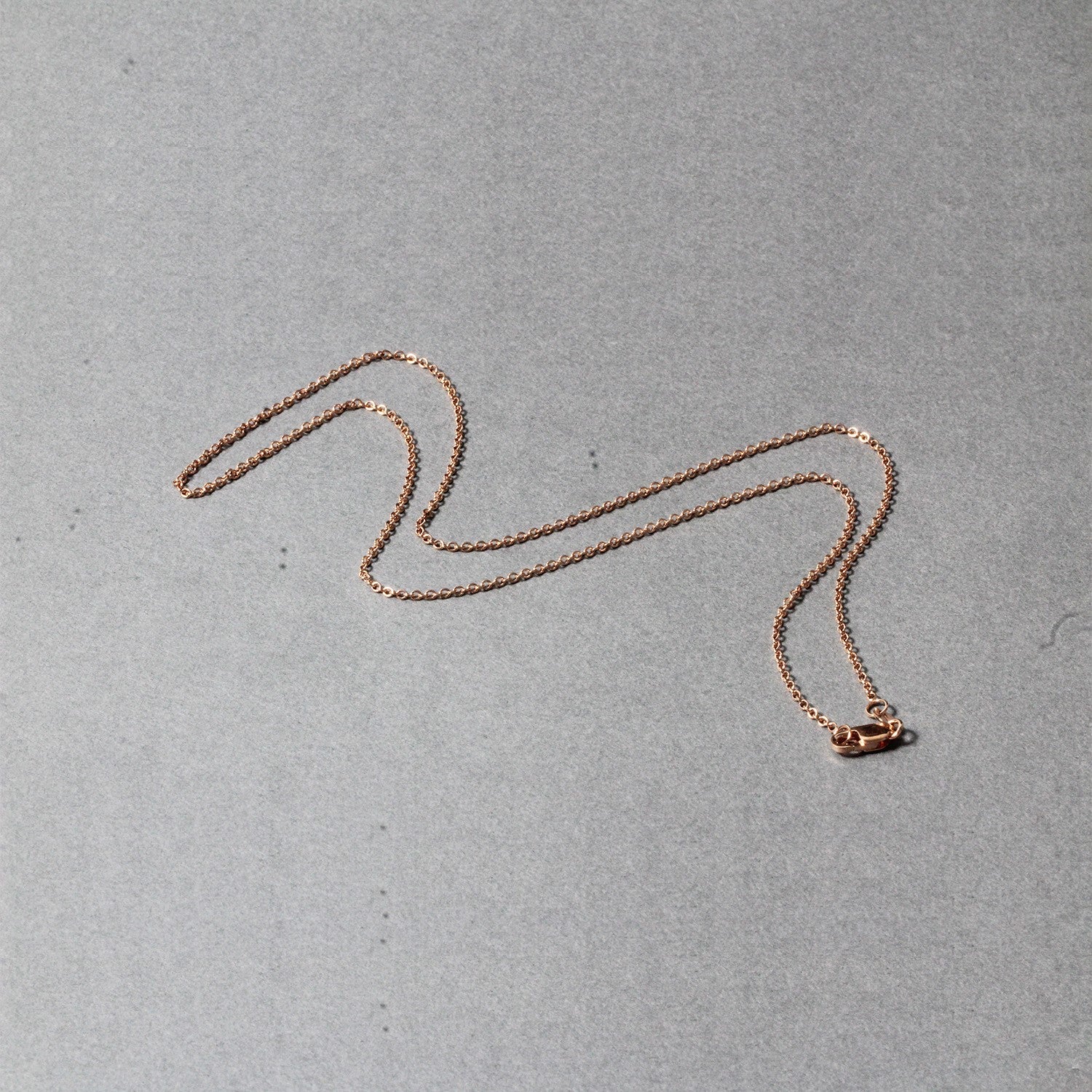 A 14k rose gold round cable link chain with a lobster clasp lies on a gray surface.