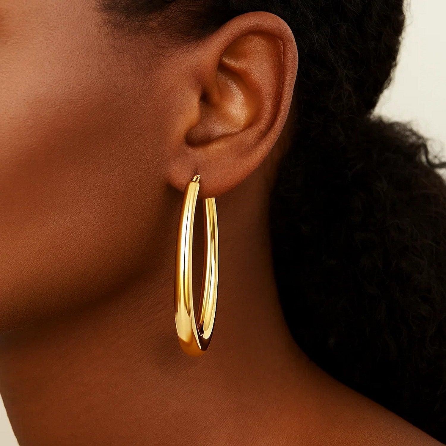 A woman wears a single 14k yellow gold elongated oval hoop earring.
