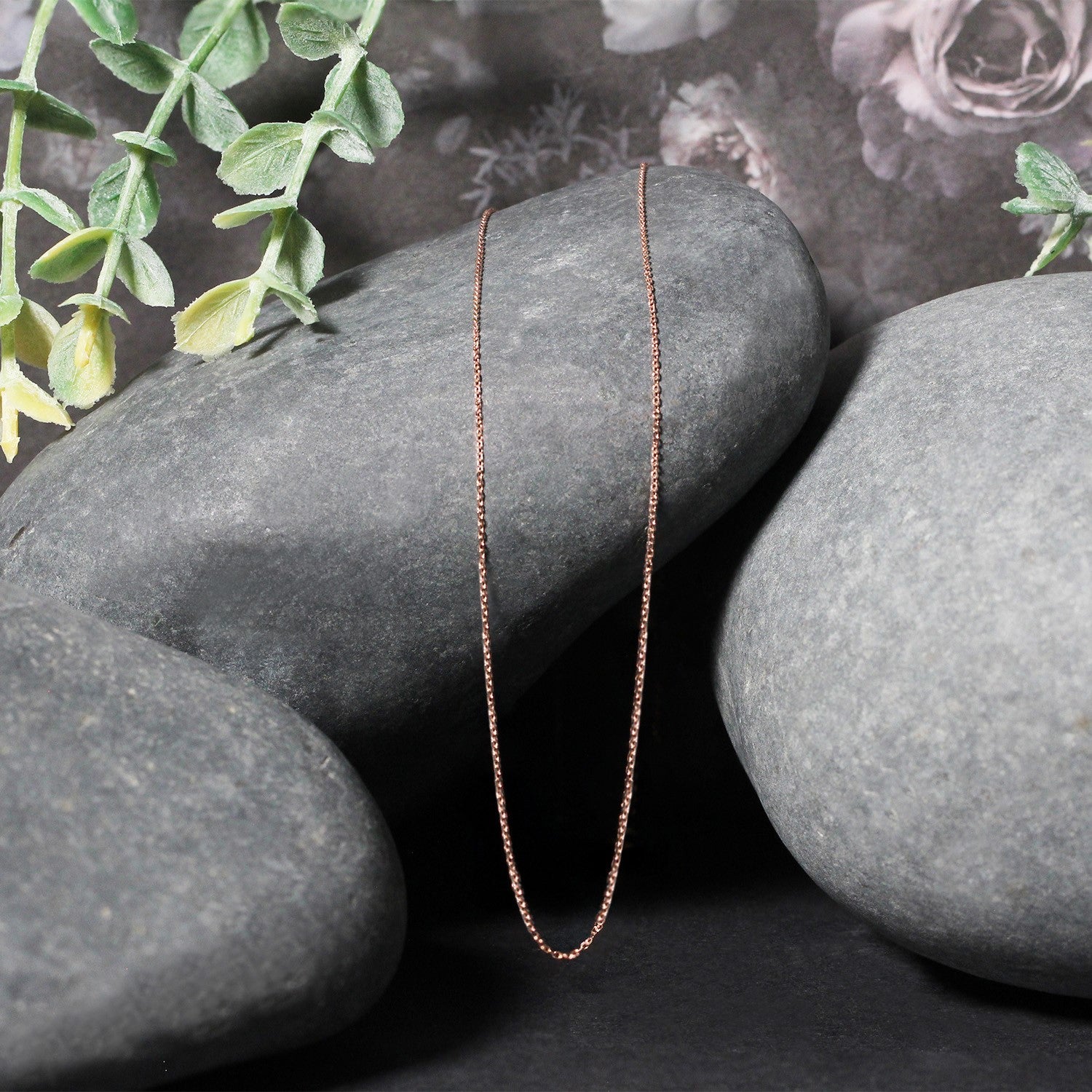 A delicate 14k rose gold diamond cut cable link chain rests on dark grey stones.