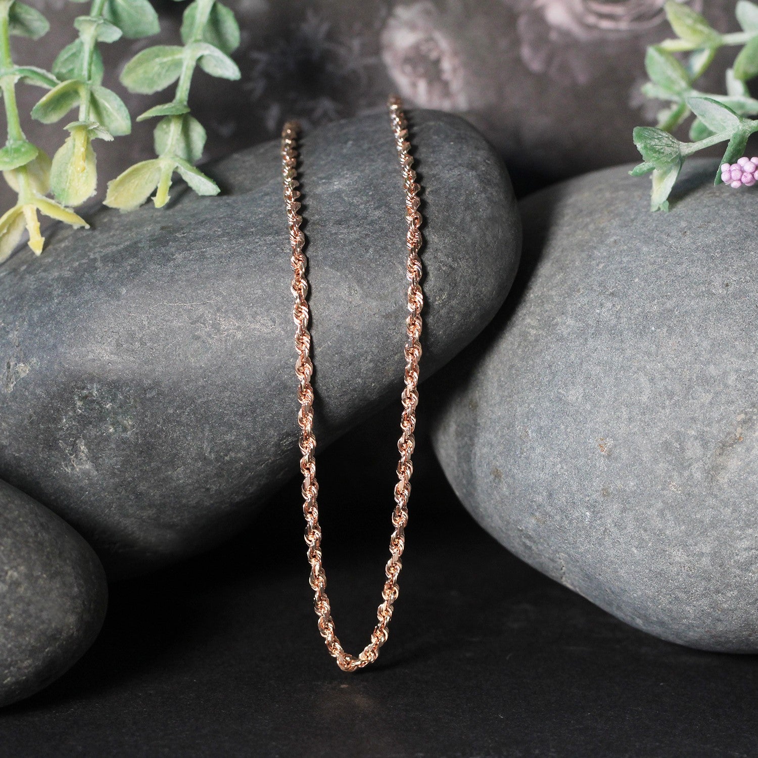 A 14k rose gold solid diamond cut rope chain, 2.30 mm wide, rests on dark rocks.