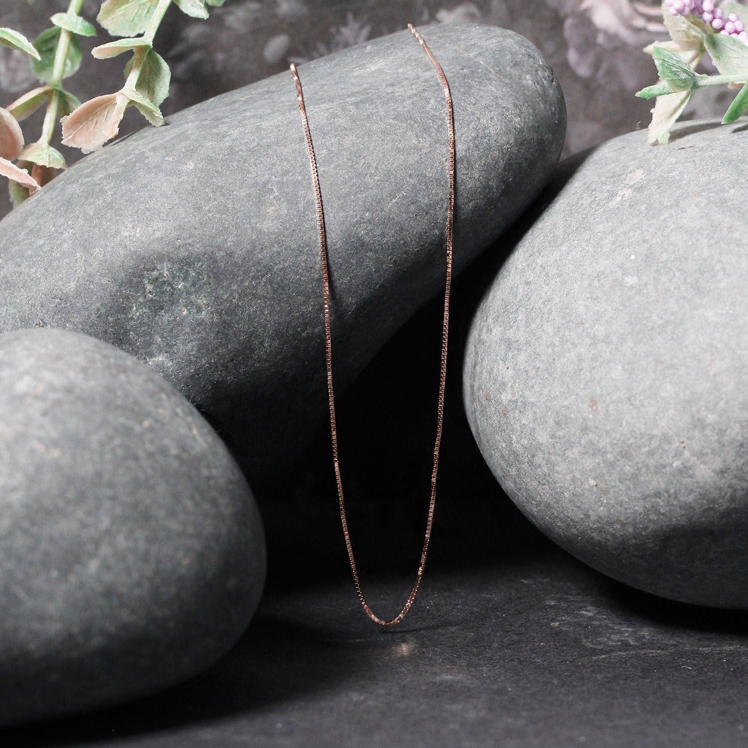 A delicate 14k rose gold box chain draped over smooth grey stones.