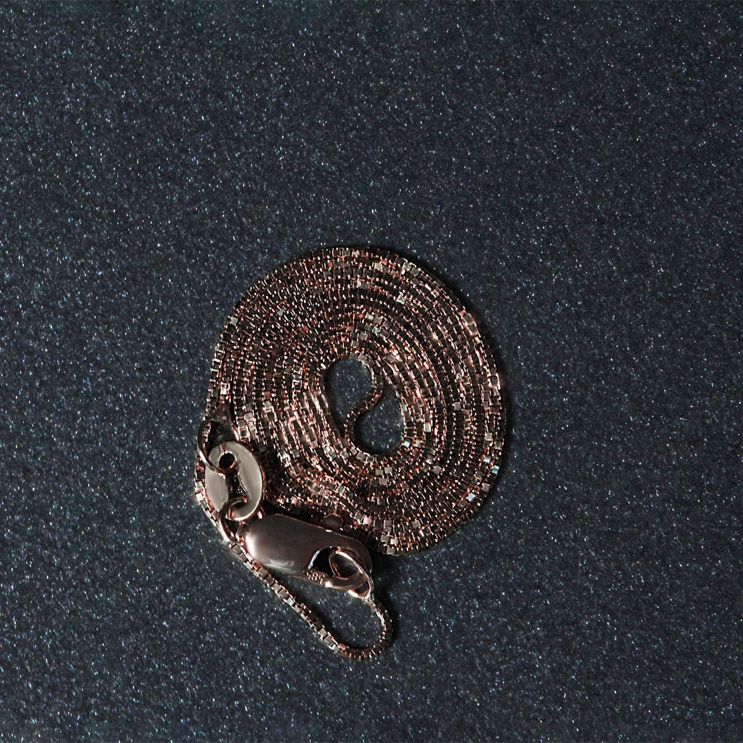 A coiled 14k rose gold box chain with a lobster clasp on a dark, glittery surface.