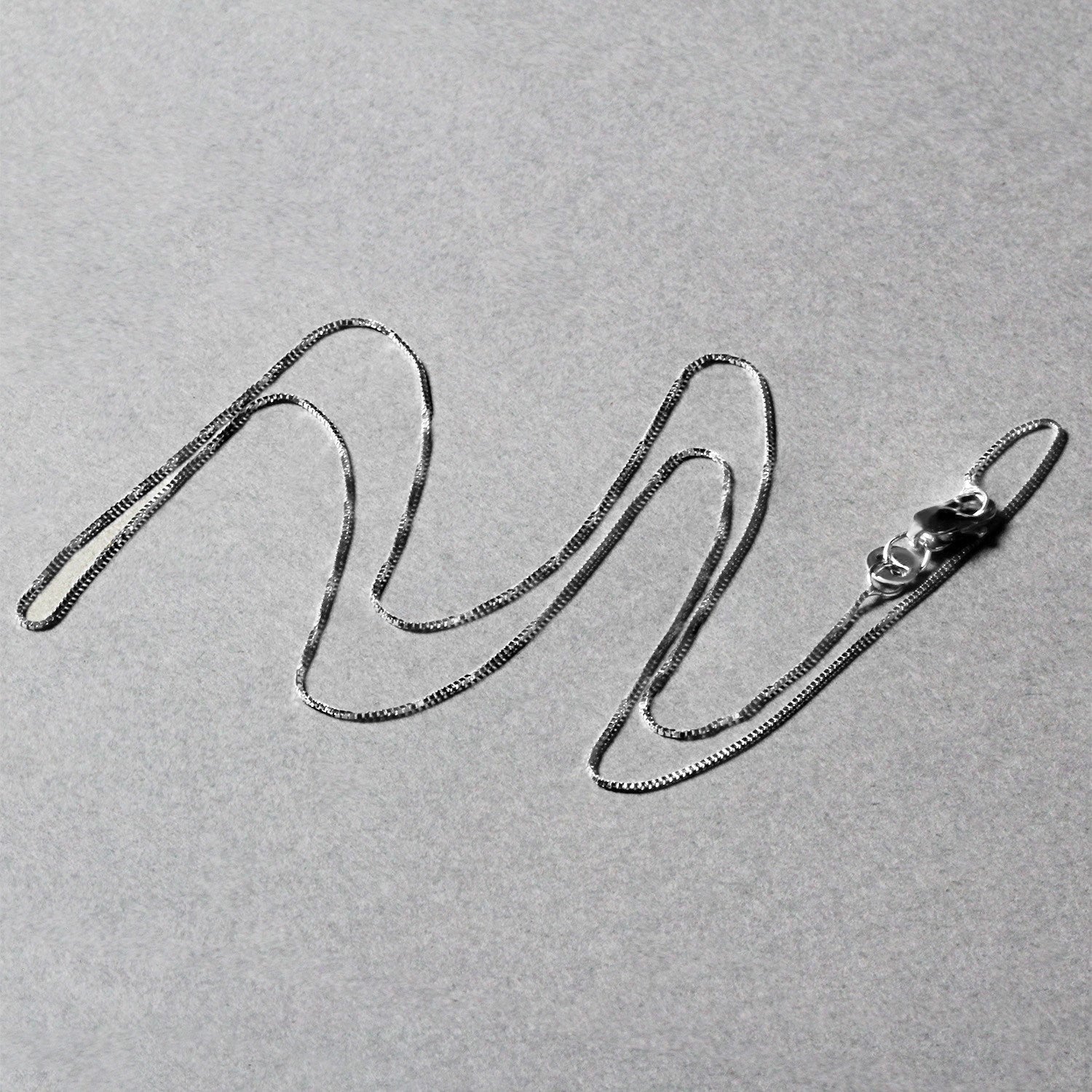 A thin, shiny white gold box chain with a lobster clasp lies on a gray surface.