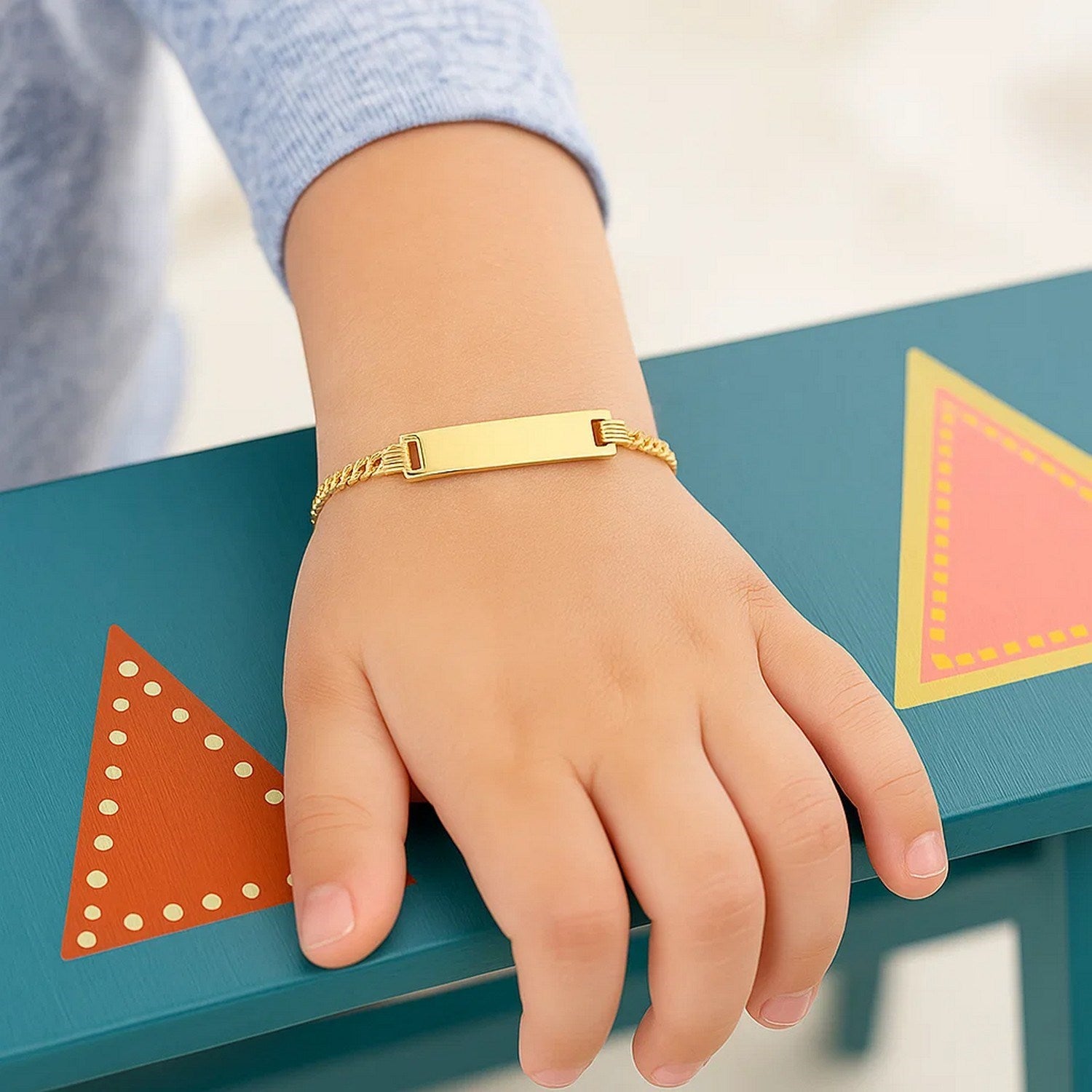 A child's hand rests on a blue surface, wearing a 14k yellow gold curb link ID bracelet.