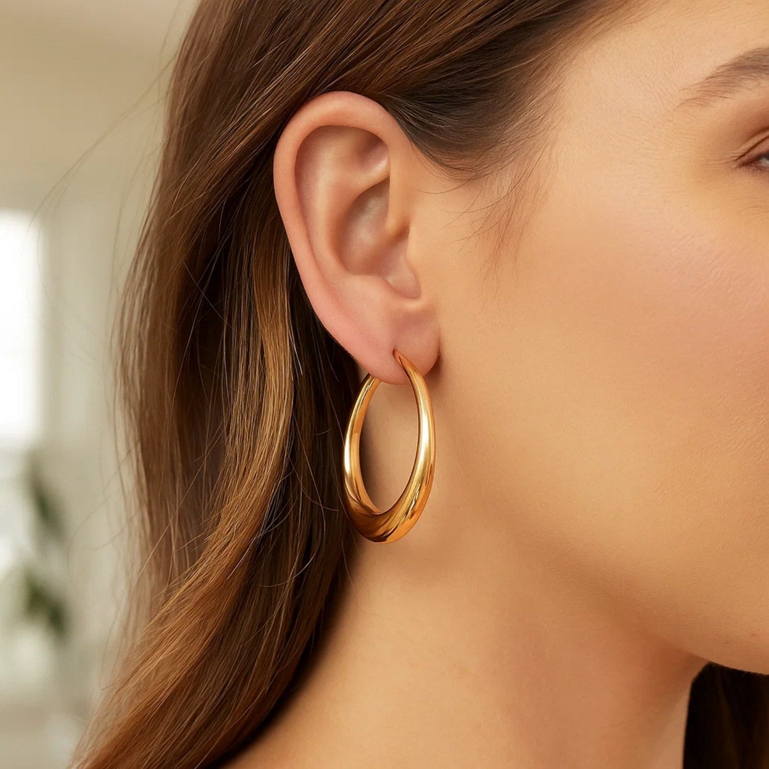 A woman wears a single polished 14K yellow gold oval hoop earring.