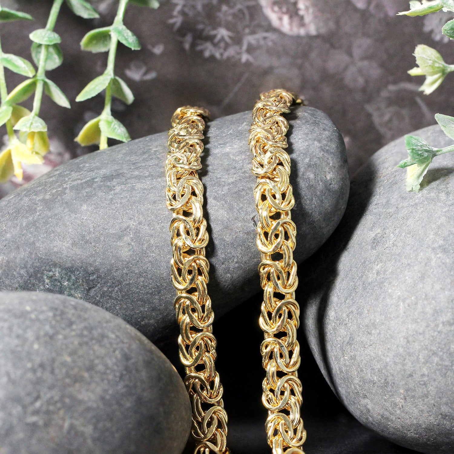 Two 14k yellow gold Byzantine chain necklaces rest on smooth grey stones.