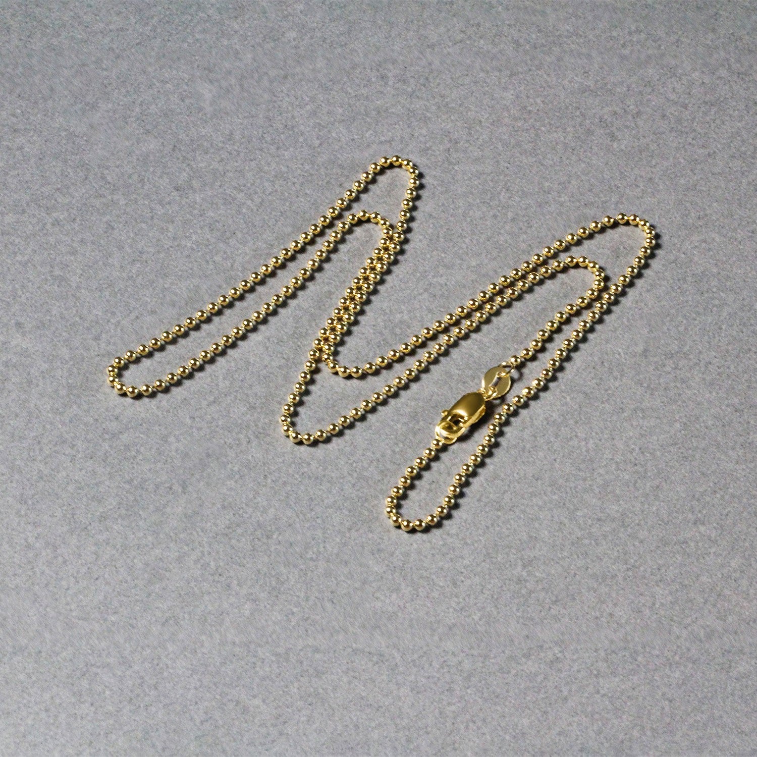 A 14k yellow gold bead chain with a lobster clasp lies on a gray surface.