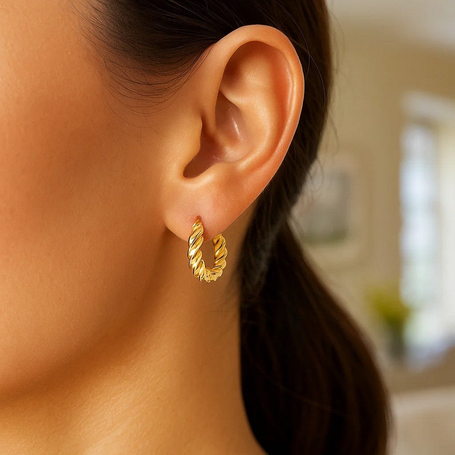 A close-up of a woman's ear wearing a polished twisted gold hoop earring.