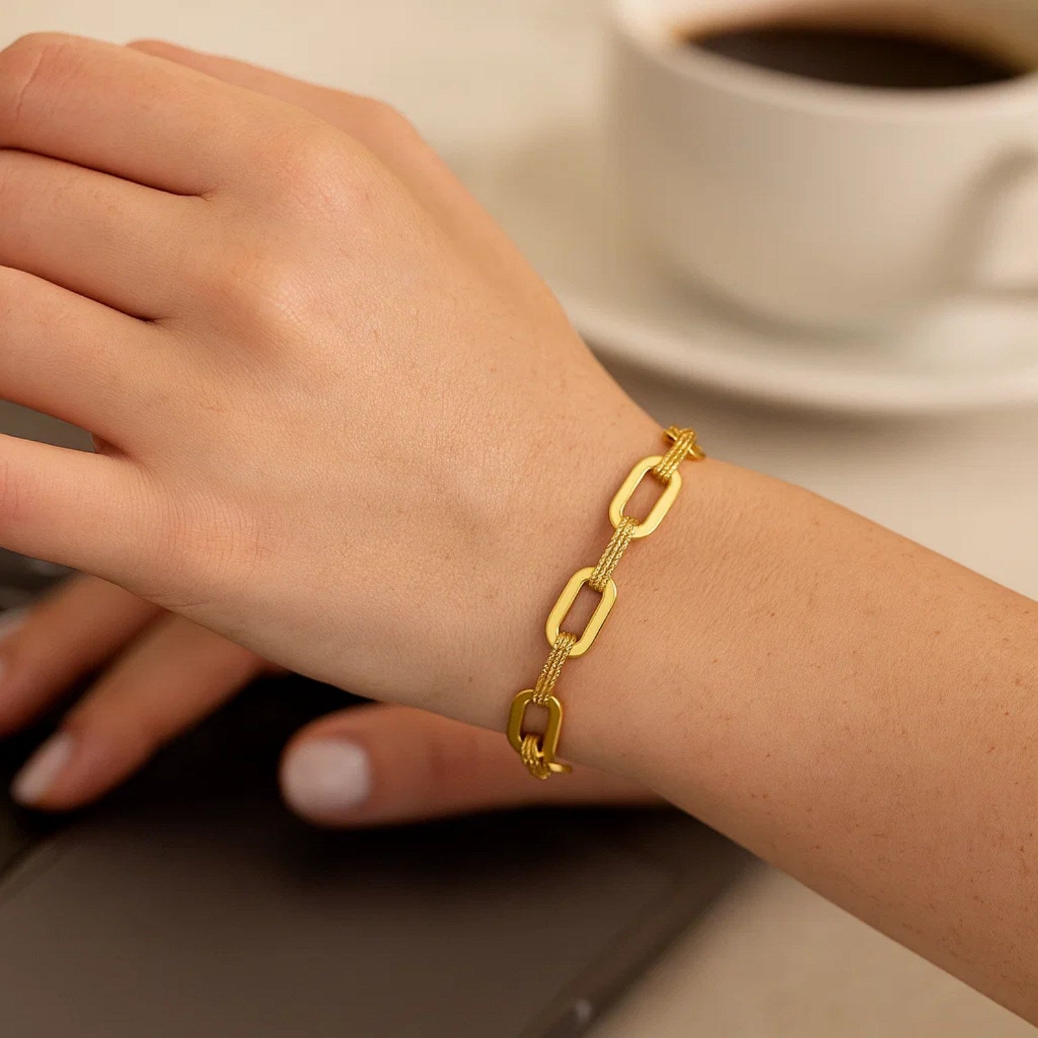 14k yellow gold paperclip chain bracelet on a woman's wrist with a coffee cup in the background.