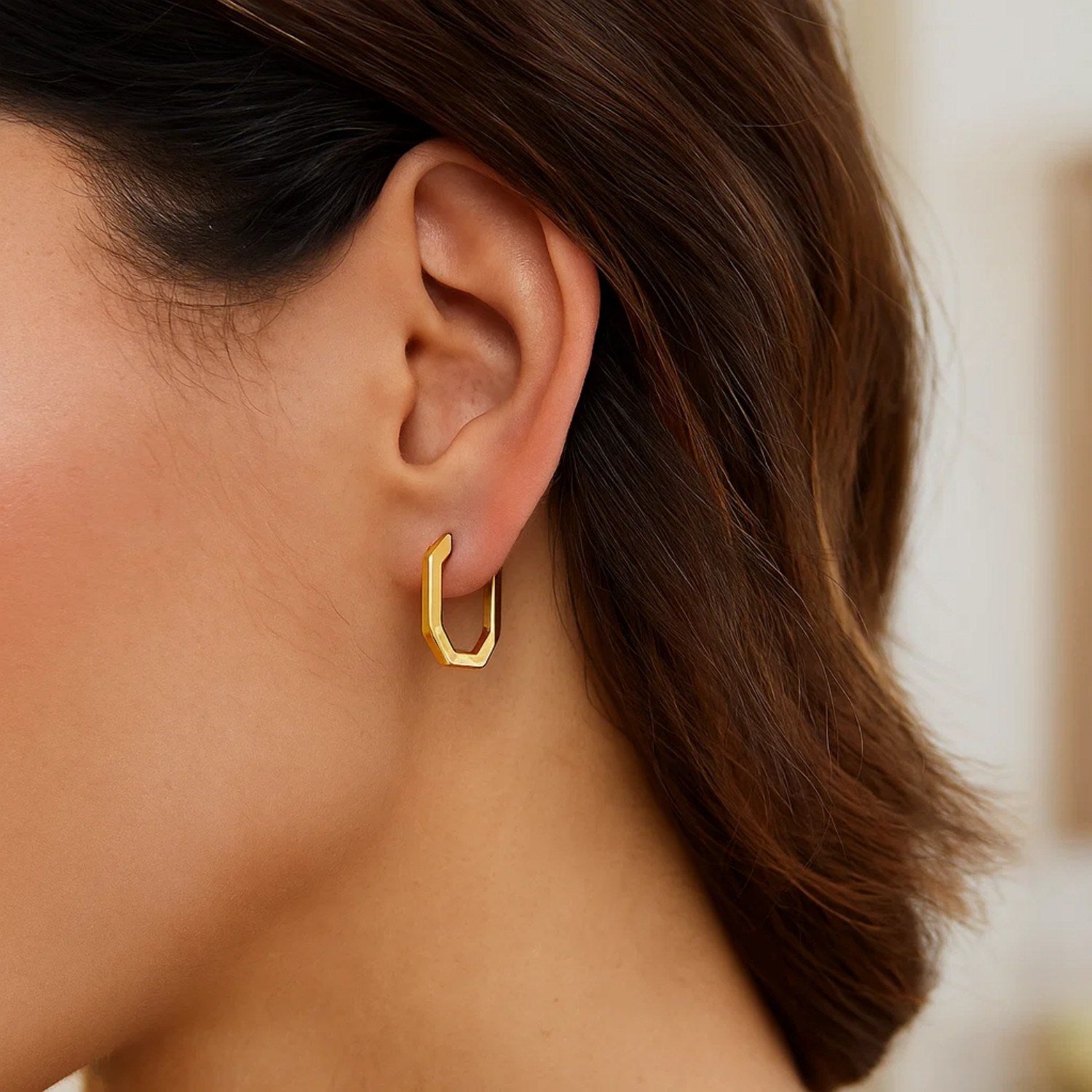 A woman wears a single 14K yellow gold hexagon hoop earring.