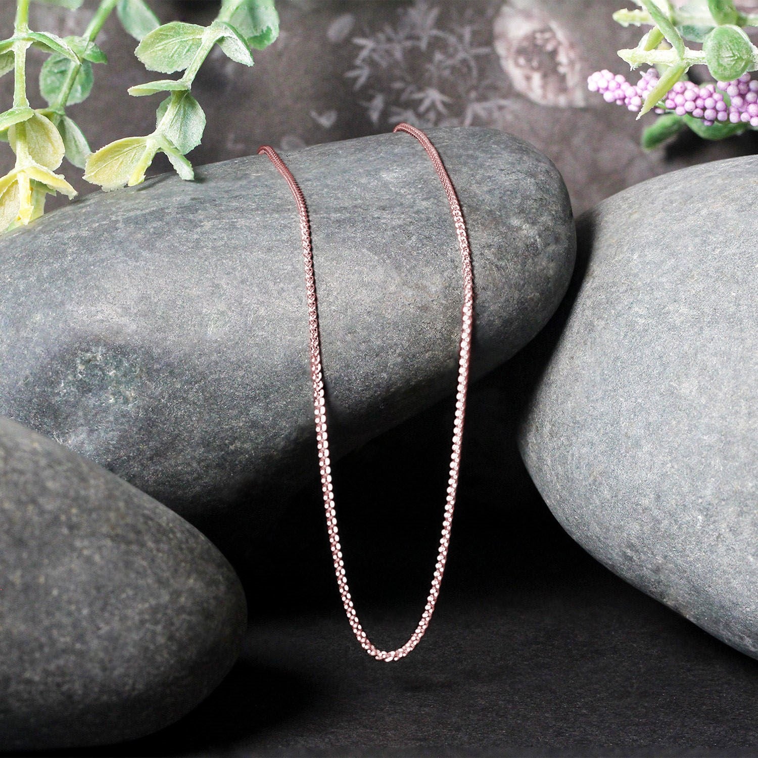 14k rose gold round wheat chain draped over dark grey stones with green and purple plants.
