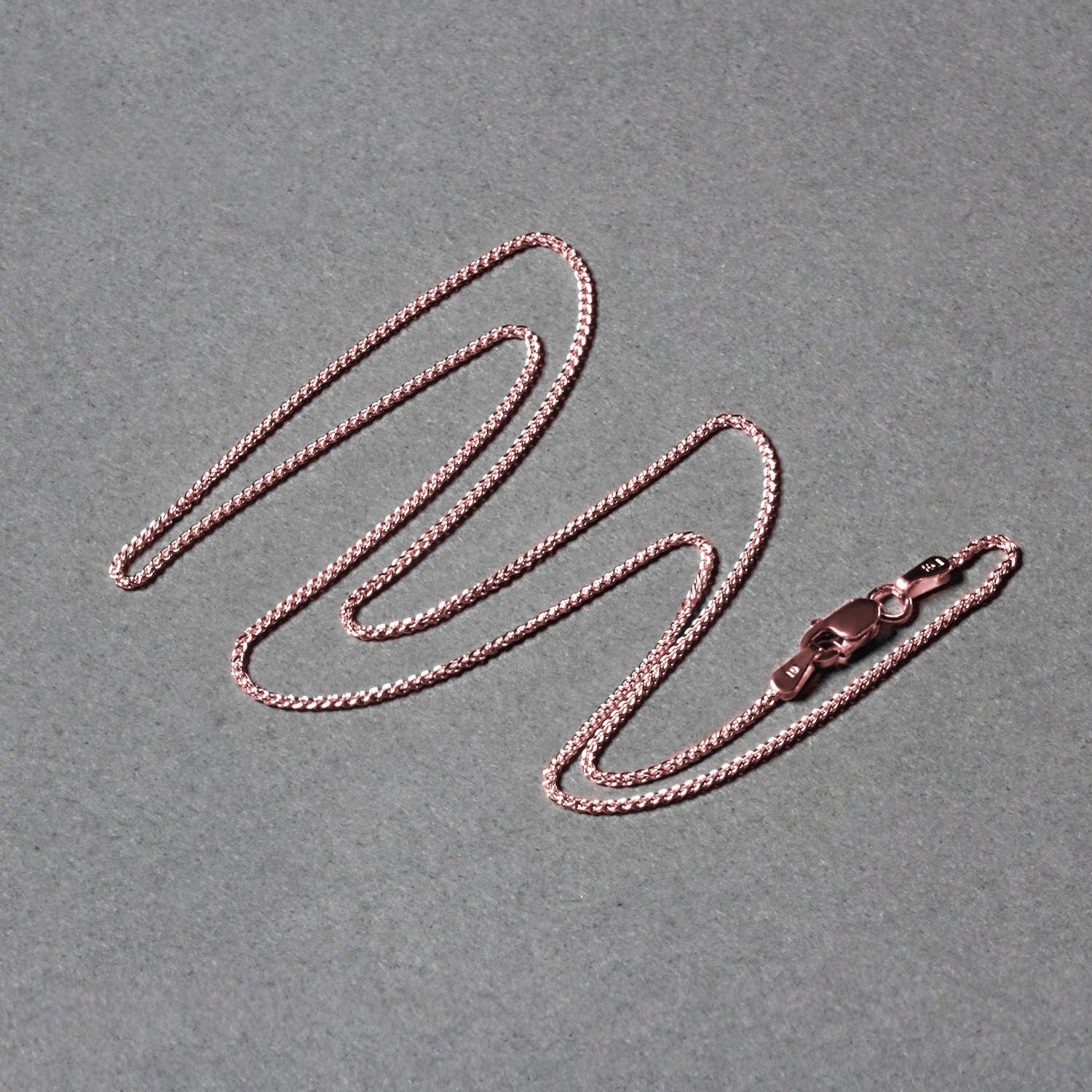 A 14k rose gold round wheat chain with a lobster clasp lies on a gray surface.