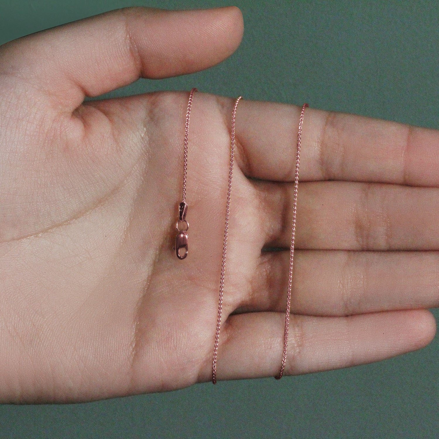 A 14k rose gold round wheat chain with a lobster clasp is displayed on a hand.
