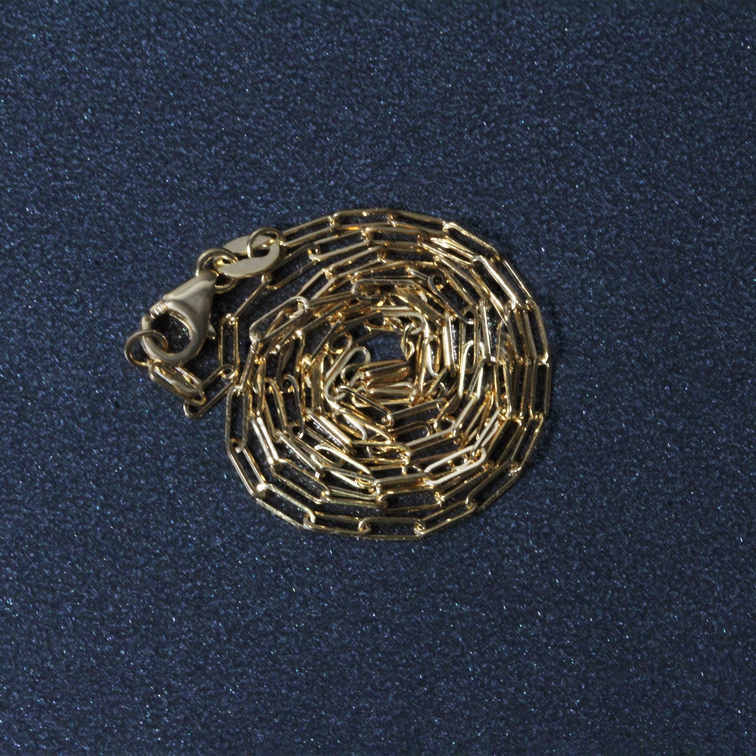 18K yellow gold paperclip chain with a lobster clasp, coiled on a dark blue surface.