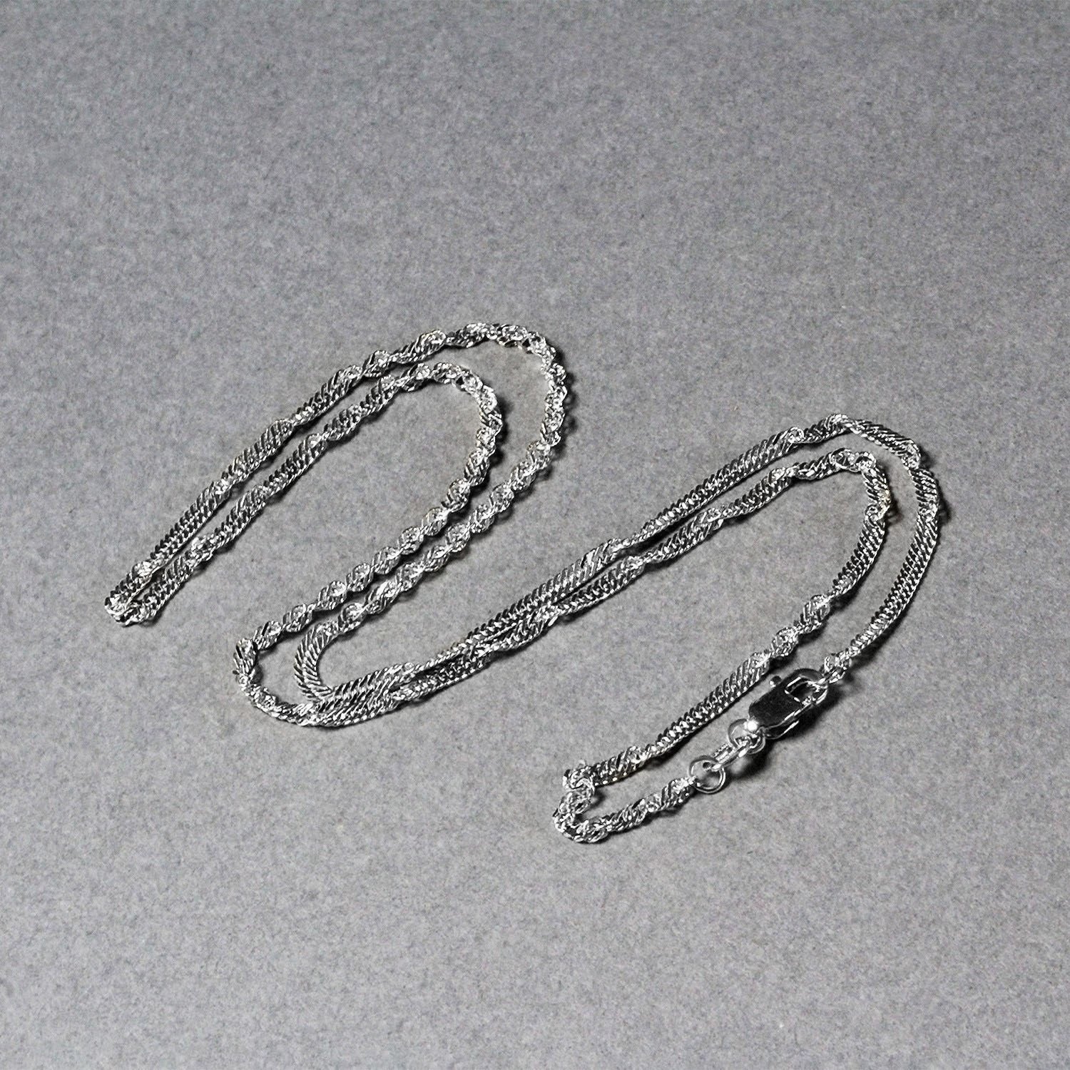 A 14k white gold Singapore chain with a lobster clasp lies on a gray surface.