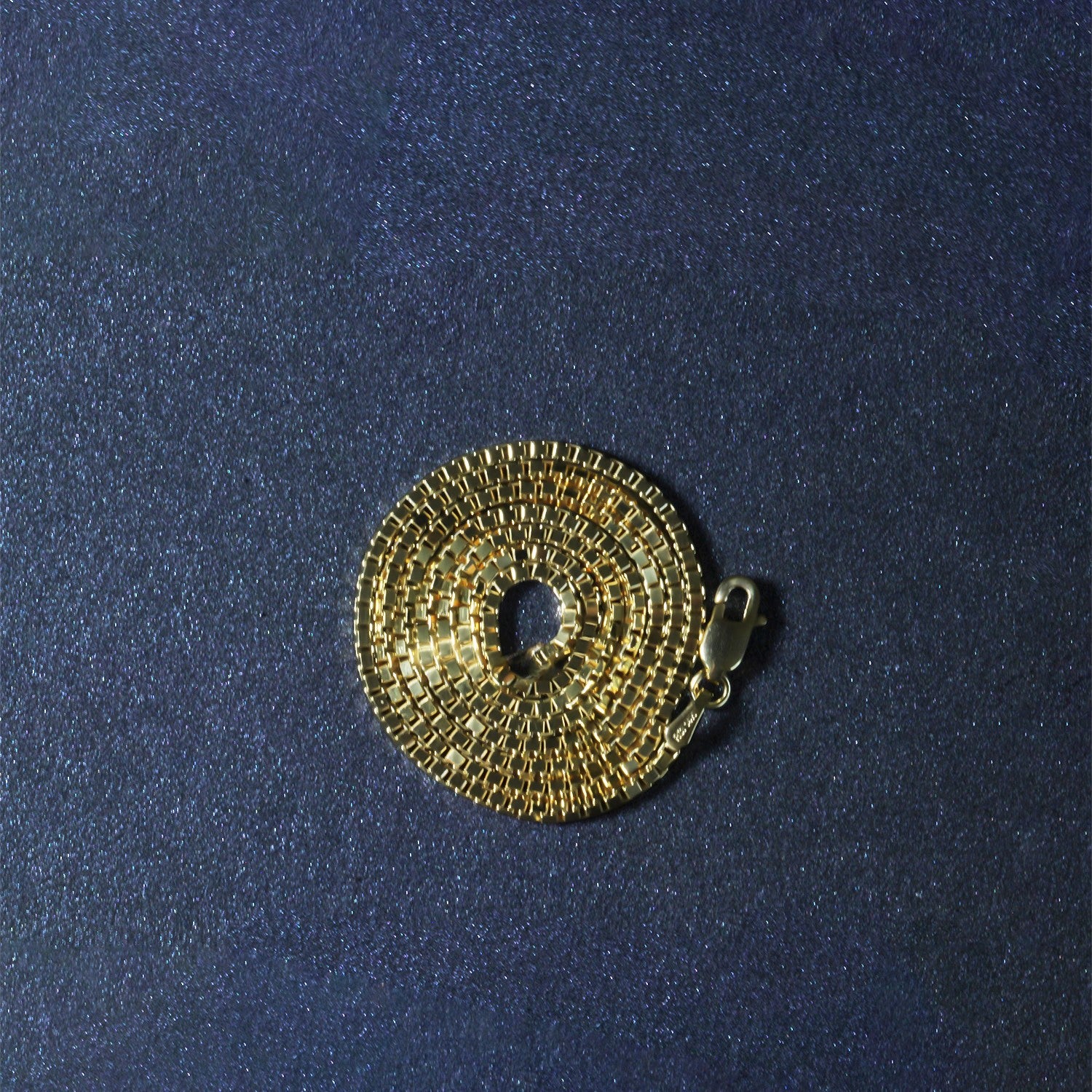 An 18k yellow gold classic box chain with a lobster clasp, coiled on a dark blue surface.