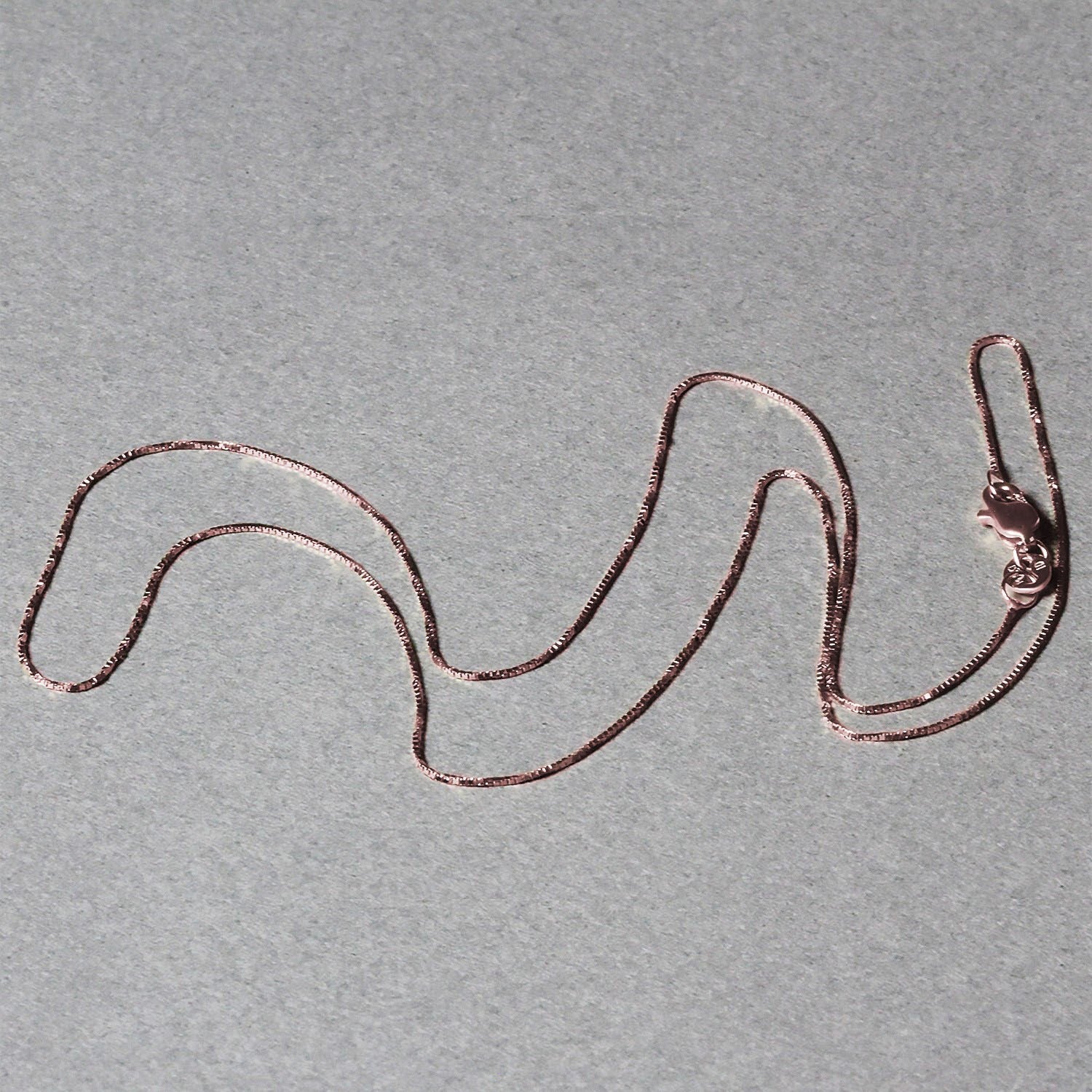 18k rose gold classic box chain with a lobster clasp, laid out on a gray surface.