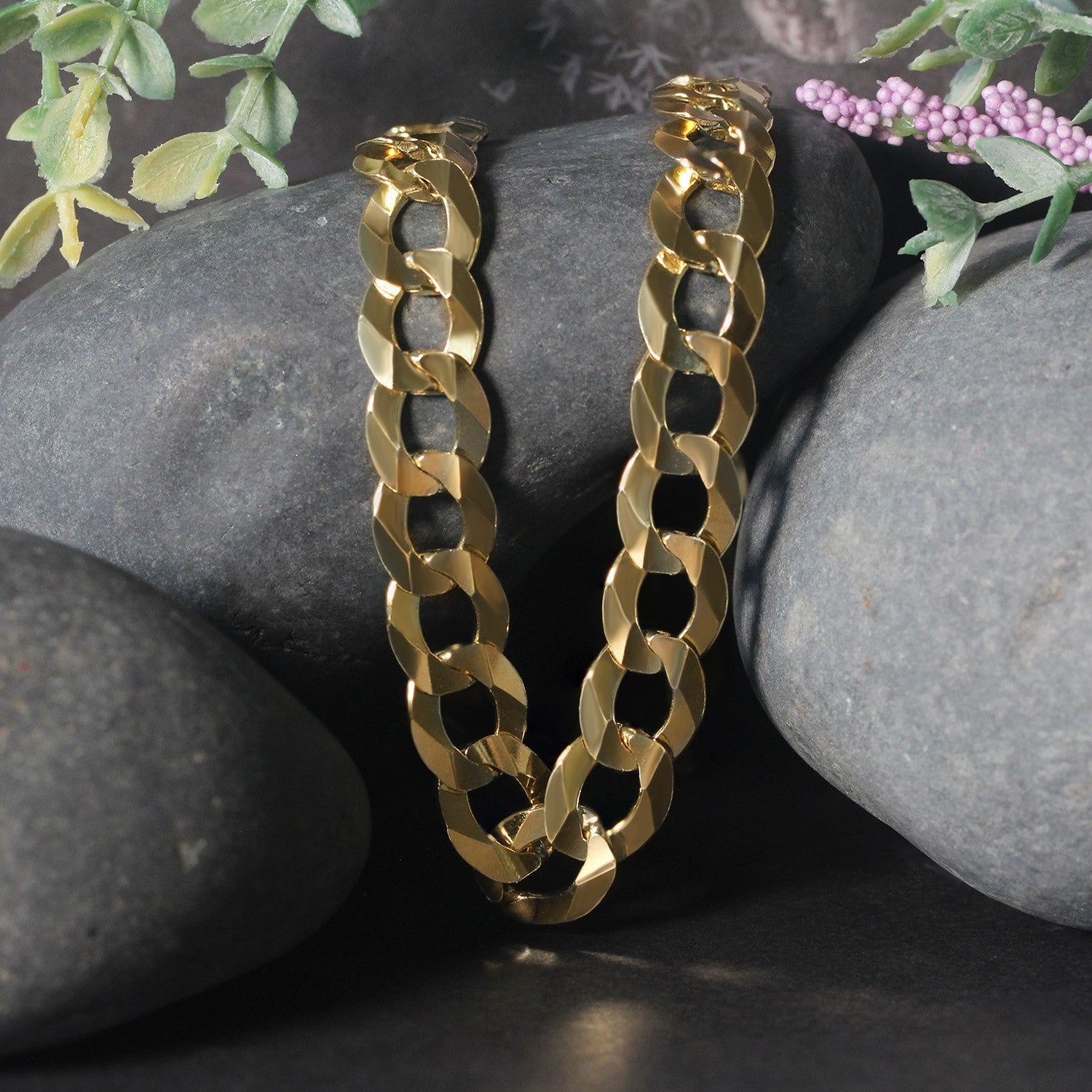 A solid 14k yellow gold curb chain rests on dark grey rocks with green and purple foliage.