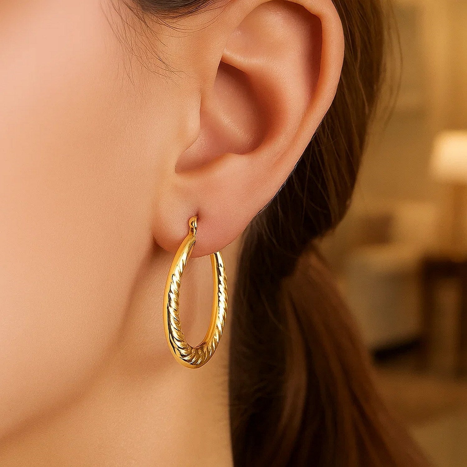 14k yellow gold oval hoop earrings with textured details on a model's ear.