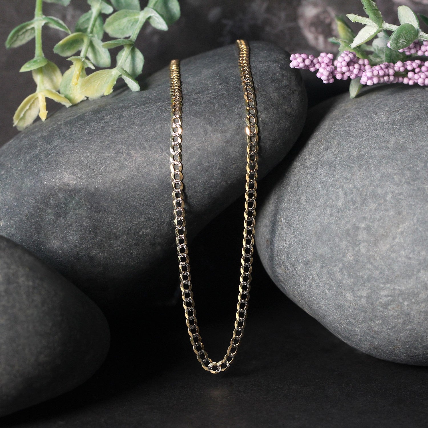 A 14k two-tone gold pave curb chain with a lobster clasp rests on dark rocks.