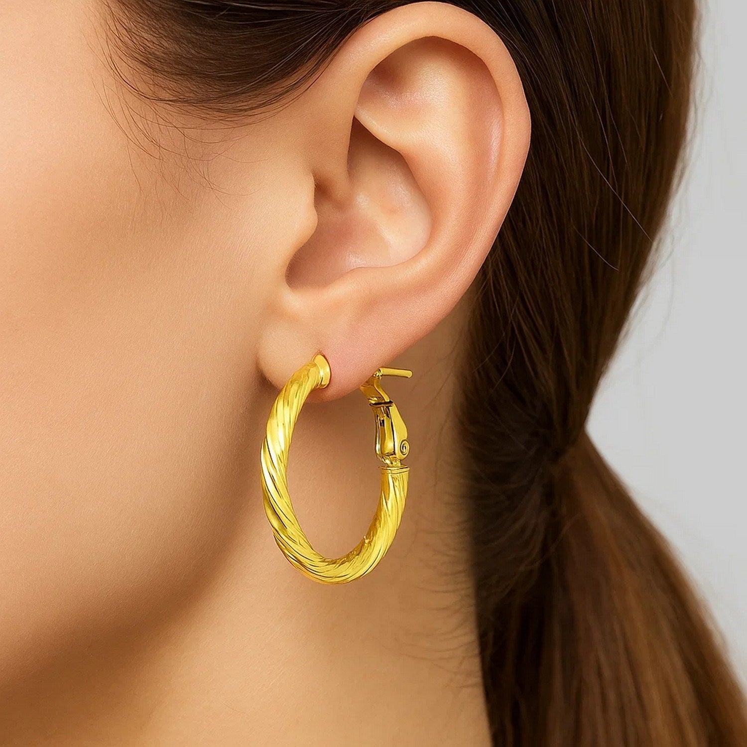 A woman wears a single twisted 14k yellow gold hoop earring with a snap clasp.
