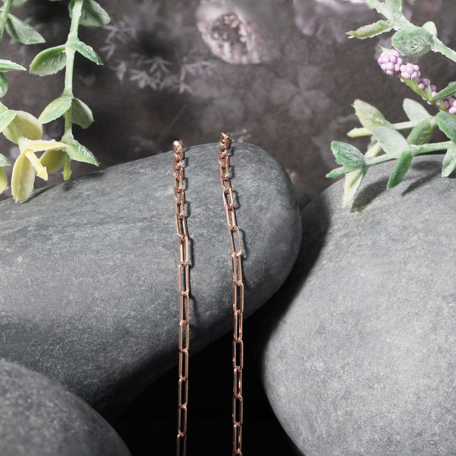 Two rose gold paperclip chains hang vertically over smooth gray stones with green foliage.