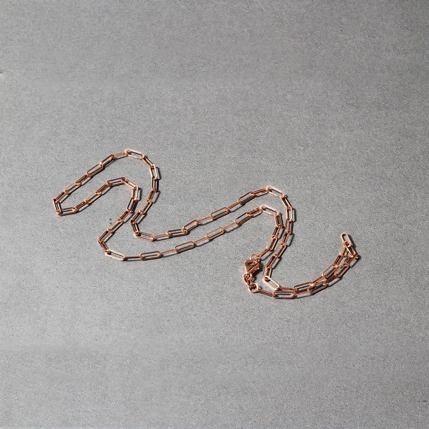 A rose gold paperclip chain with a lobster clasp lies on a gray surface.