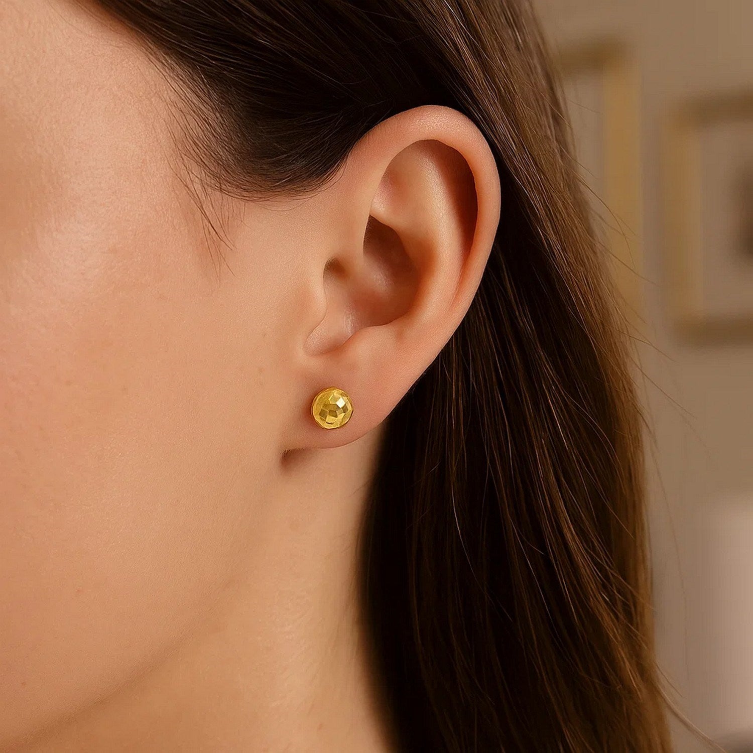A single 14k yellow gold faceted ball earring is worn on a woman's ear.