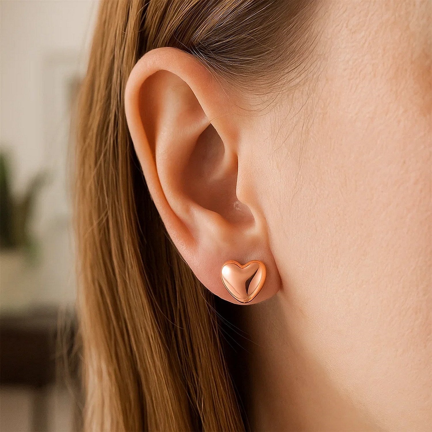A rose gold puffed heart earring is worn on a person's ear.