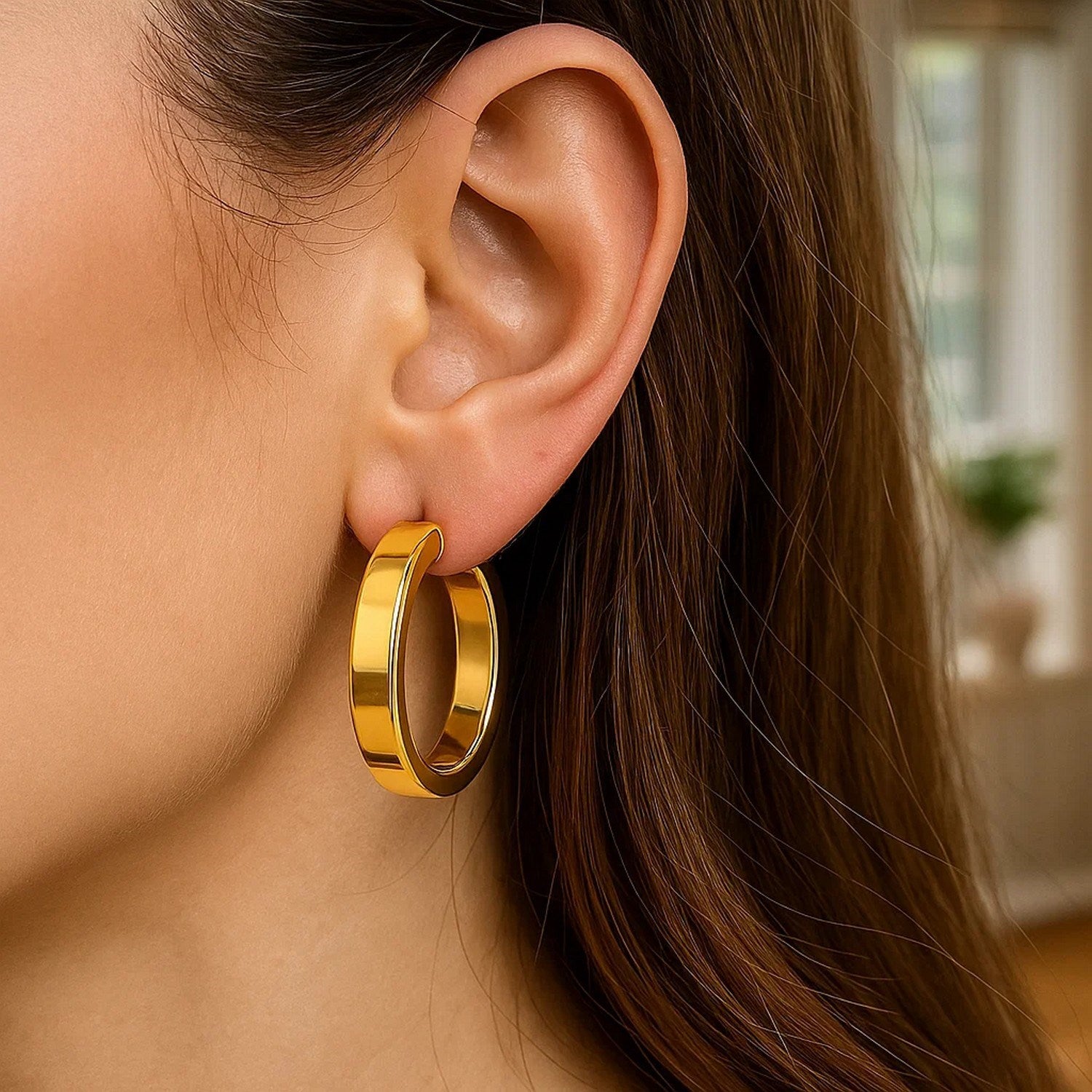Large tube-shaped hoop earrings in 14K yellow gold are worn on a woman's ear.